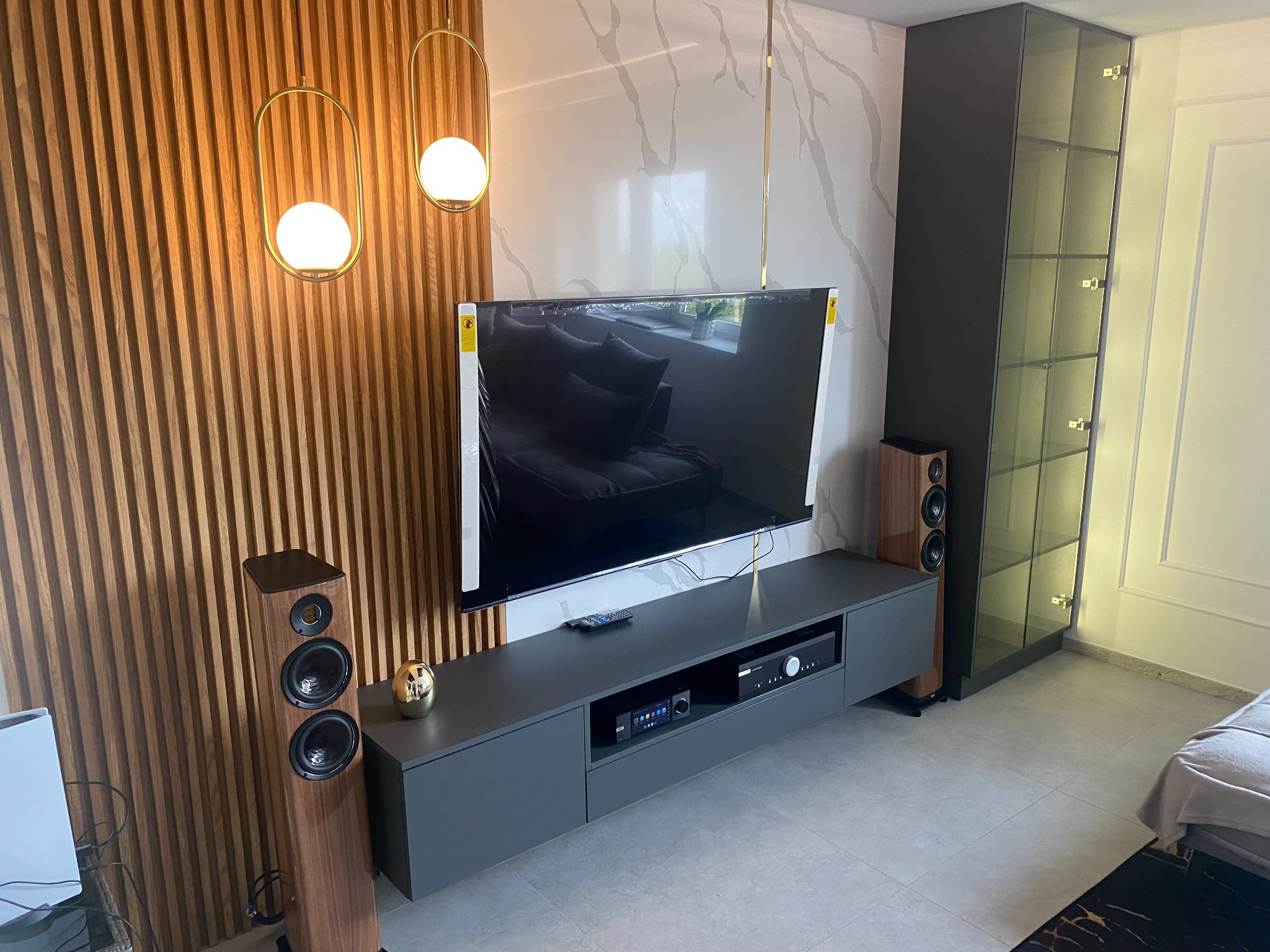 Nowoczesny salon z drewnianą ścianą lamelową, szarą szafką RTV z wbudowanym sprzętem audio, głośnikami podłogowymi i wiszącymi lampami kulami, uzupełniony o przeszkloną szafę.