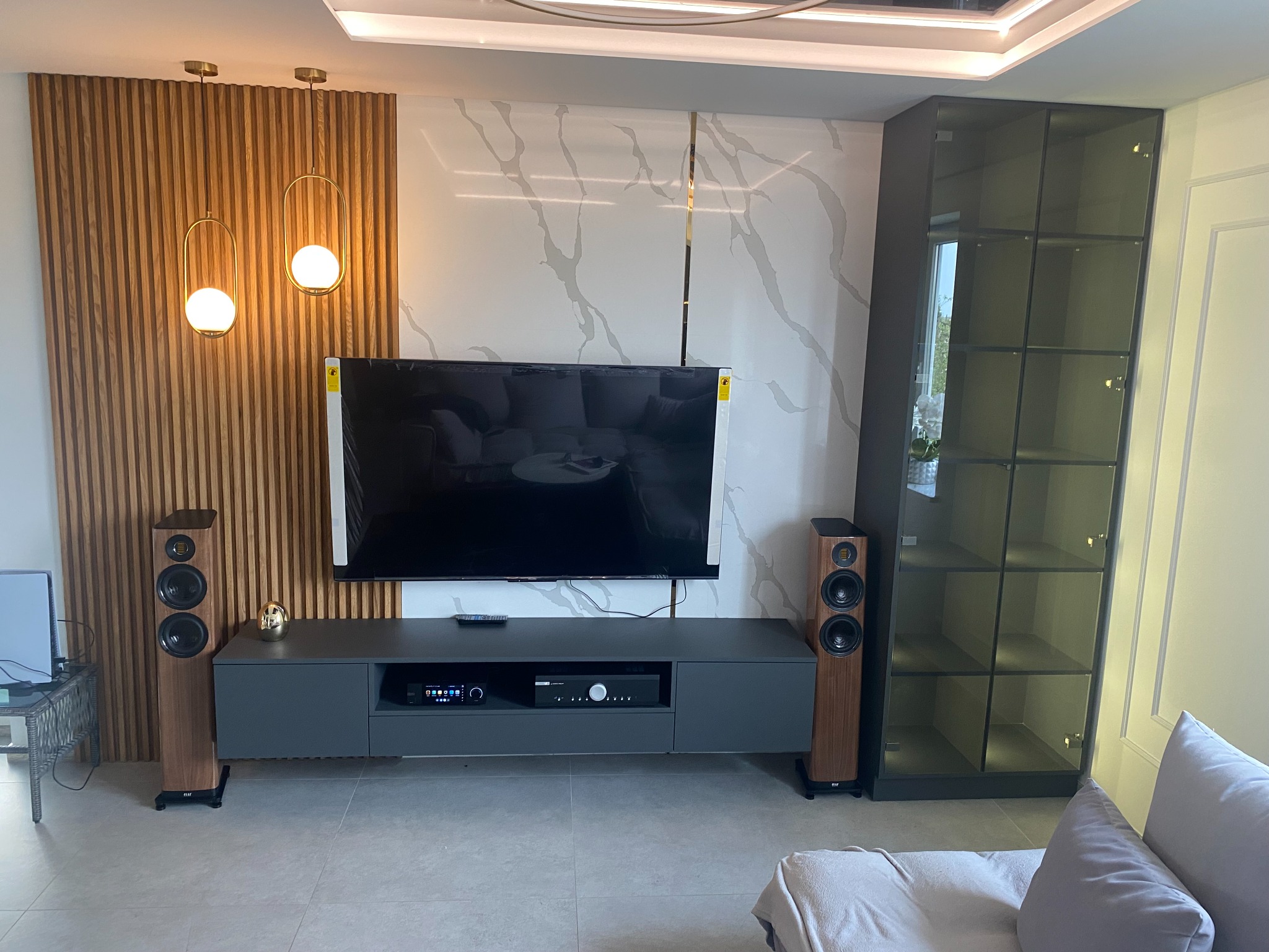 Nowoczesny salon z szarą szafką RTV, drewnianą ścianą z lamelami, marmurową ścianą z złotymi wstawkami, szarą witryną i drewnianymi głośnikami podłogowymi.