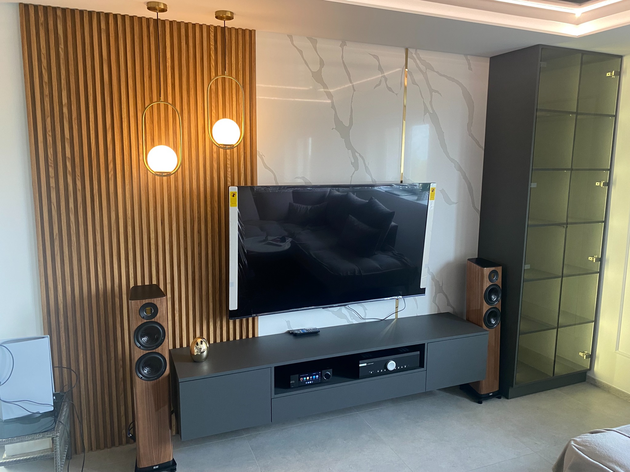 Nowoczesny salon z drewnianą ścianą, marmurowym panelem, szarą szafką RTV, głośnikami podłogowymi i przeszkloną szafą, oświetlony złotymi lampami wiszącymi.