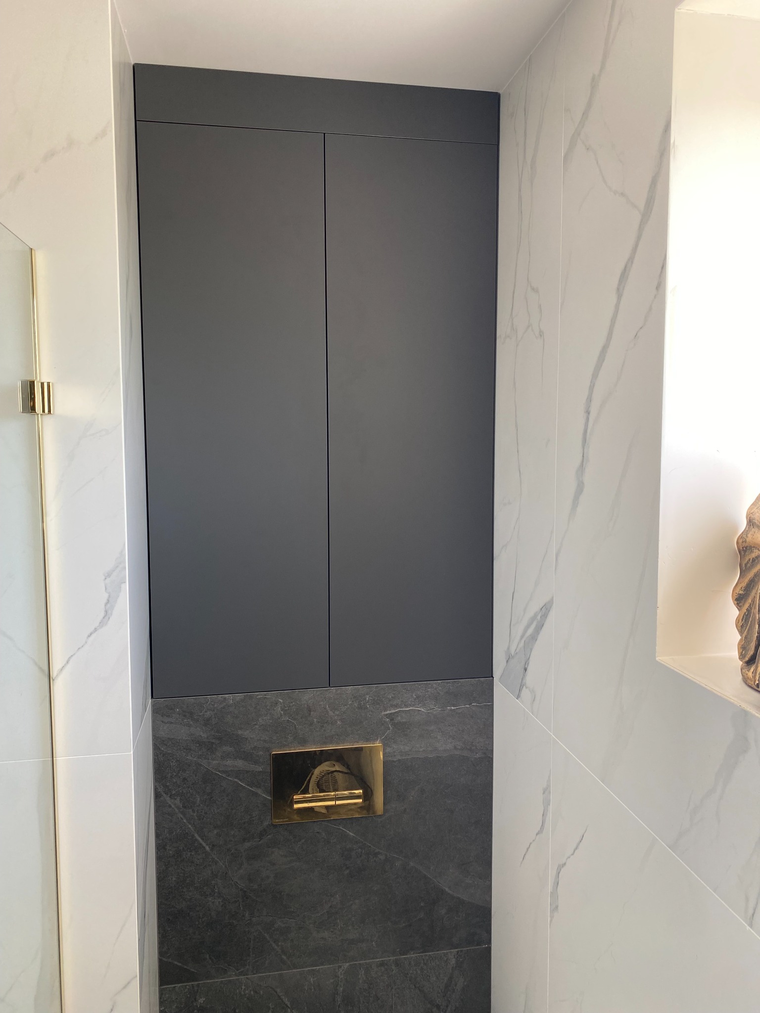 Szara szafka wnękowa zlicowana ze ścianą z białego marmuru, nad podwieszaną toaletą z czarnym marmurem i złotym przyciskiem.
