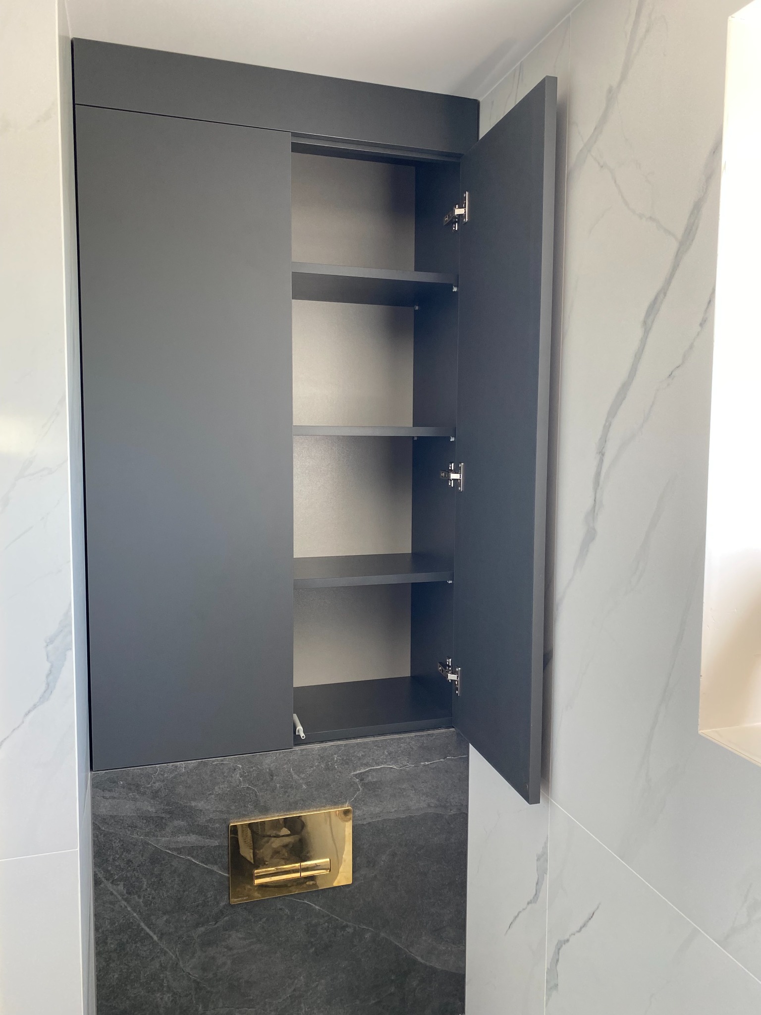 Szara szafka łazienkowa z otwartymi drzwiczkami, widoczne trzy półki. Szafka umieszczona nad czarnym blatem z złotym przyciskiem spłuczki. Tło stanowią białe płytki imitujące marmur.