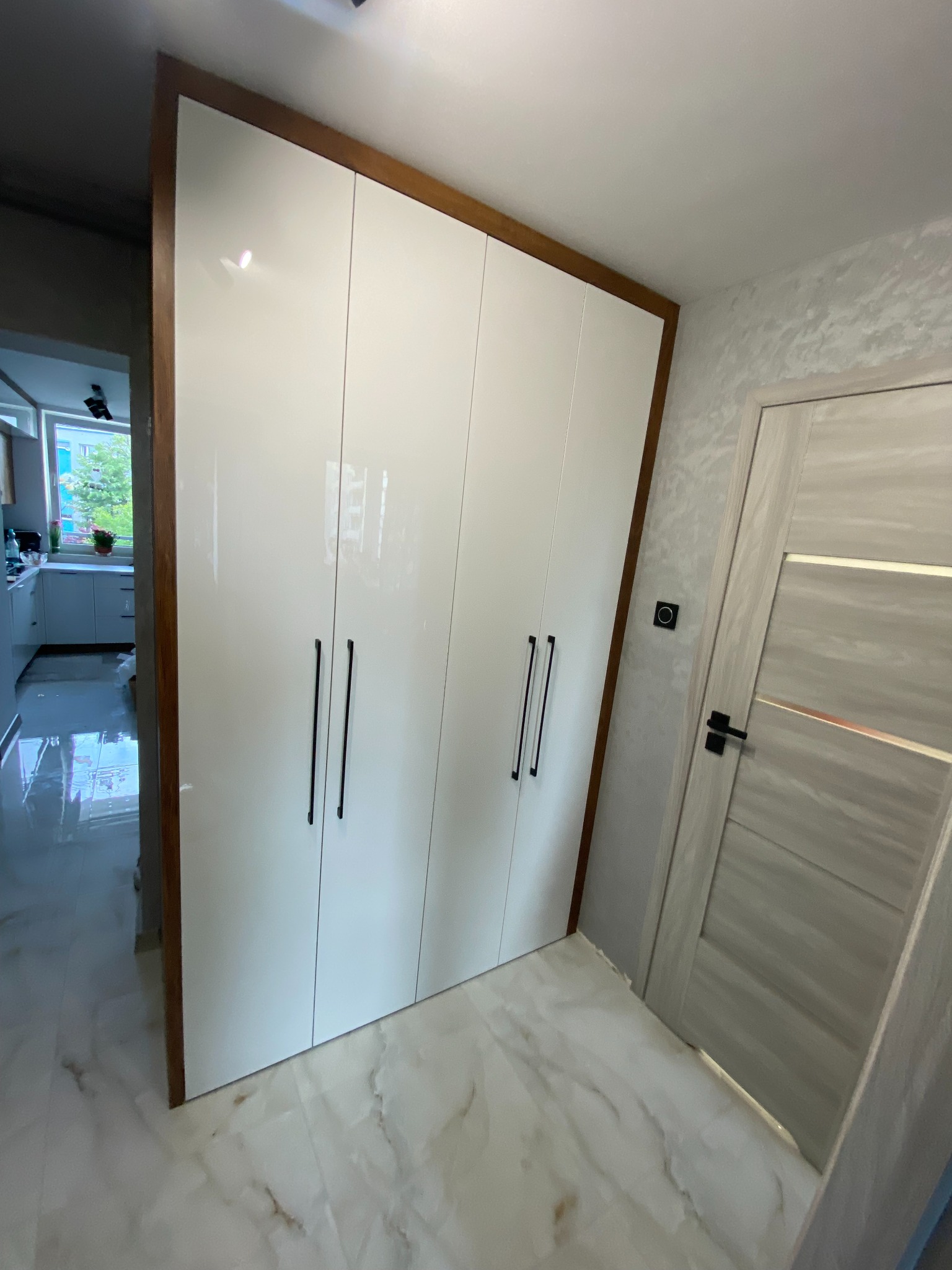 Biała, lakierowana szafa wnękowa z ciemnymi, pionowymi uchwytami, otoczona drewnianą ramą, widoczna na tle marmurowej podłogi i drzwi wewnętrznych w jasnej tonacji.