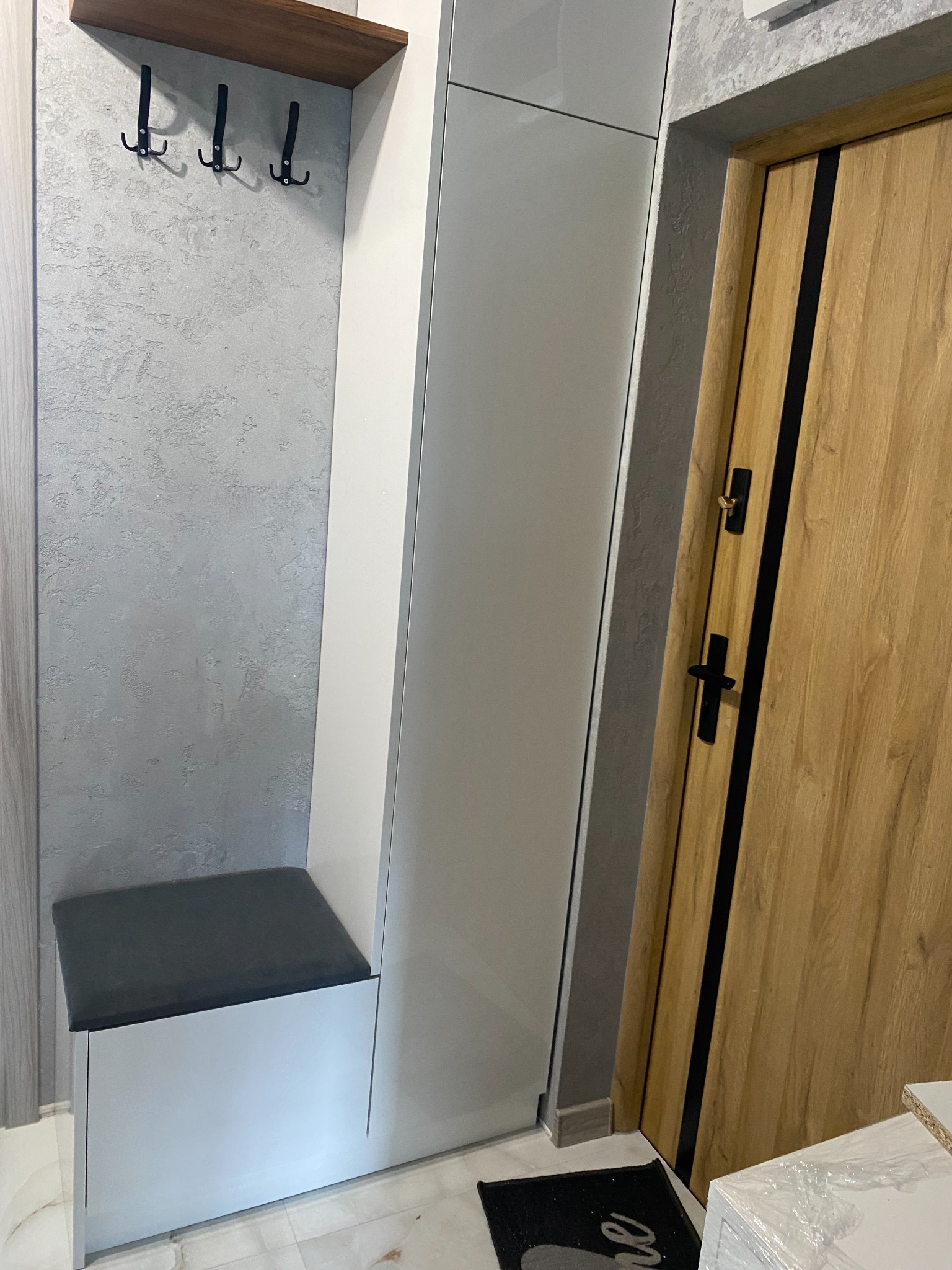 Wnętrze przedpokoju: biała zabudowa z siedziskiem, wieszakami i szafą, obok drewniane drzwi z czarnymi akcentami, szara ściana o fakturze tynku.