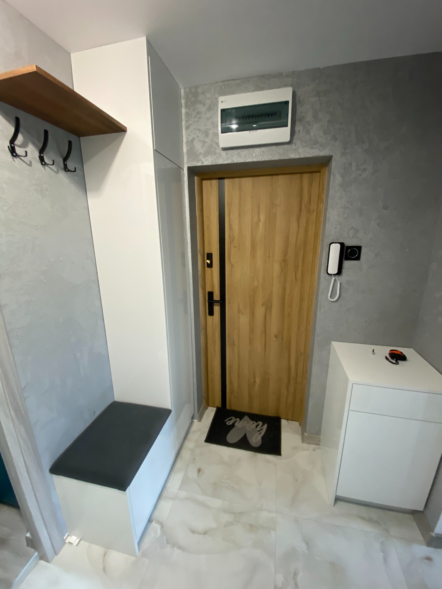 Wnętrze przedpokoju z zabudową: biała szafa z siedziskiem, drewniana półka z wieszakami, drzwi wejściowe z drewnianym fornirem i czarnymi akcentami, biała komoda z domofonem na ścianie w tle.