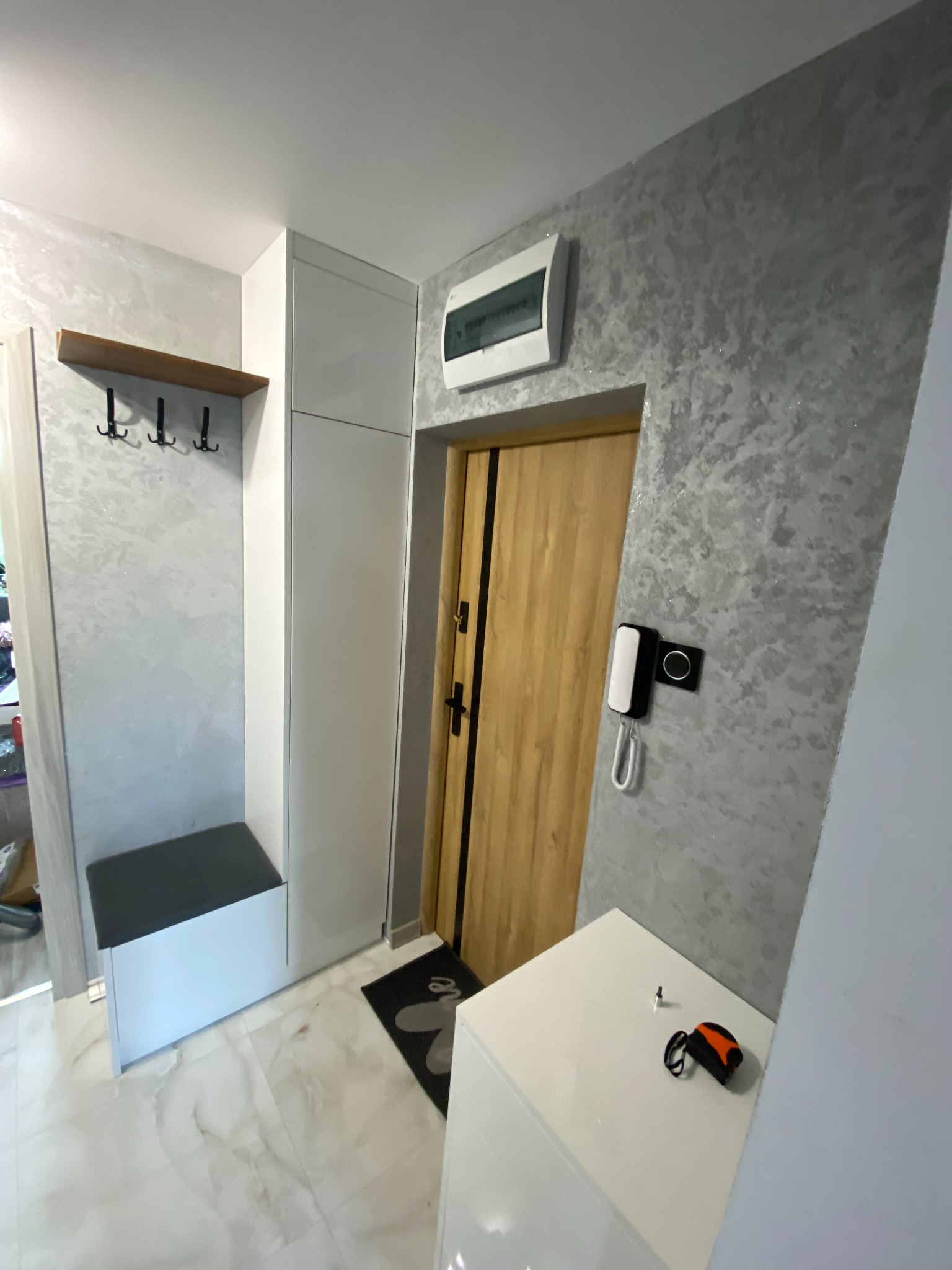 Przedpokój z szafą na wymiar w zabudowie, siedziskiem i wieszakami, ściany pokryte dekoracyjnym tynkiem, widoczny fragment drzwi wejściowych i miernik na szafce.