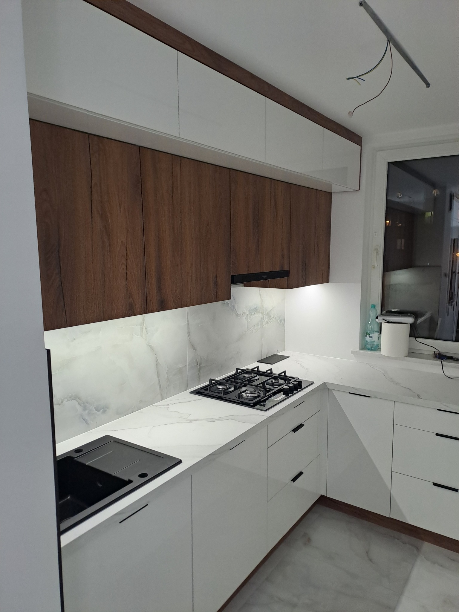Nowoczesna kuchnia z białymi szafkami dolnymi i drewnopodobnymi górnymi, marmurowy blat i backsplash, czarny zlew i płyta grzewcza, widoczne kable wystające z sufitu.