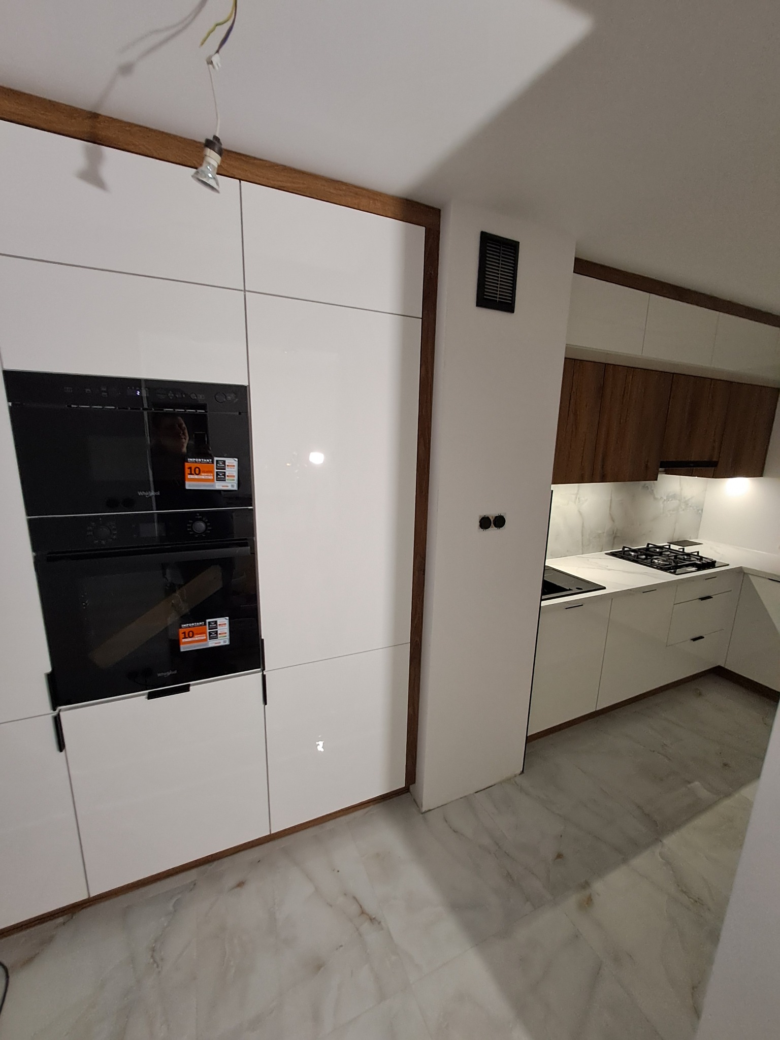 Nowoczesna biała kuchnia z zabudowanym piekarnikiem i płytą gazową, drewniane akcenty wykończeniowe, marmurowa podłoga, widoczna instalacja elektryczna lampy sufitowej.