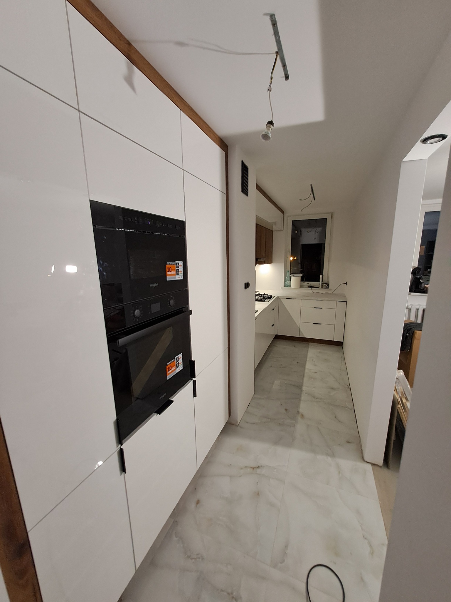 Biała zabudowa kuchenna z czarnym piekarnikiem i mikrofalówką w trakcie montażu, widoczne niedokończone oświetlenie sufitowe i marmurowe płytki podłogowe.