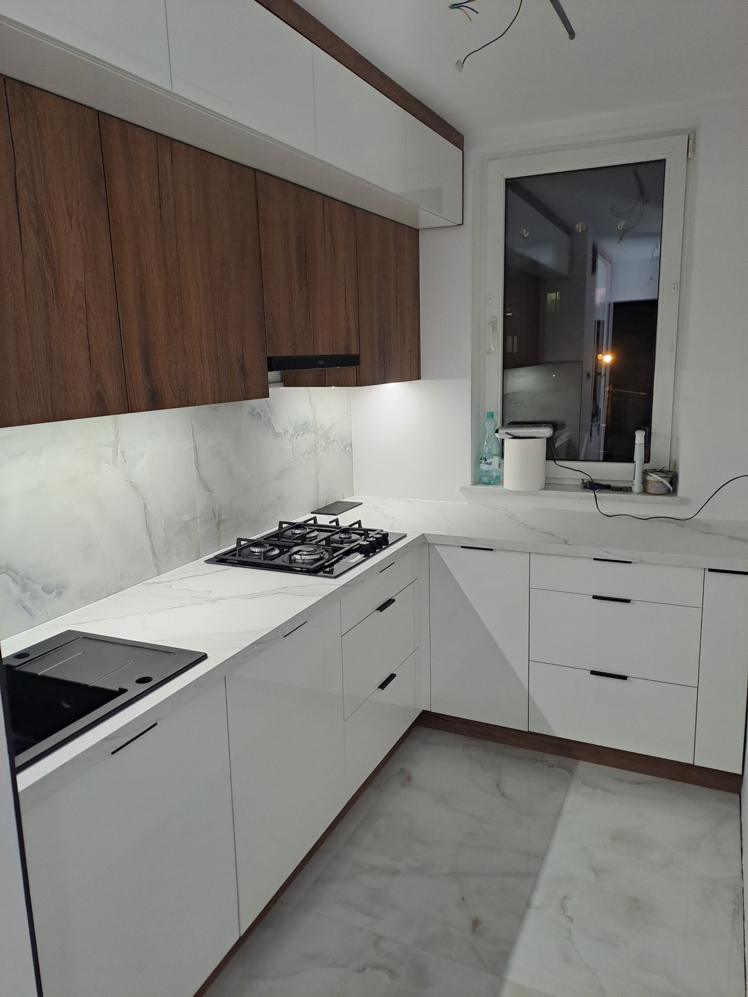 Nowoczesna kuchnia z białymi szafkami, drewnianymi górnymi szafkami i blatem imitującym marmur, czarna płyta gazowa i zlewozmywak, widoczne kable wystające z sufitu.