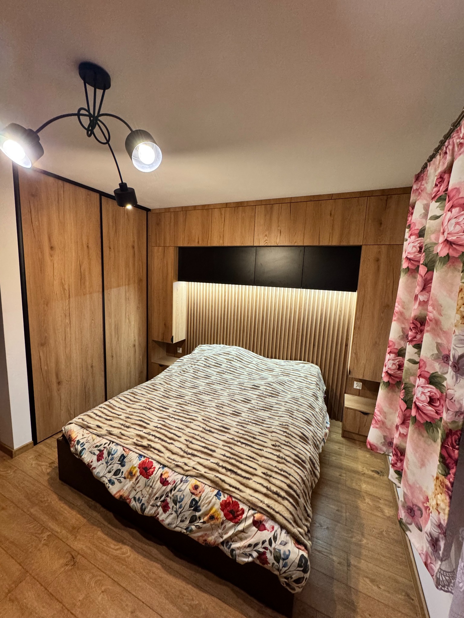 Szafa wnękowa z drewnianymi frontami i czarnymi obramowaniami, zintegrowana z zagłówkiem łóżka w sypialni. Widoczna lampa sufitowa i zasłony w kwiaty.