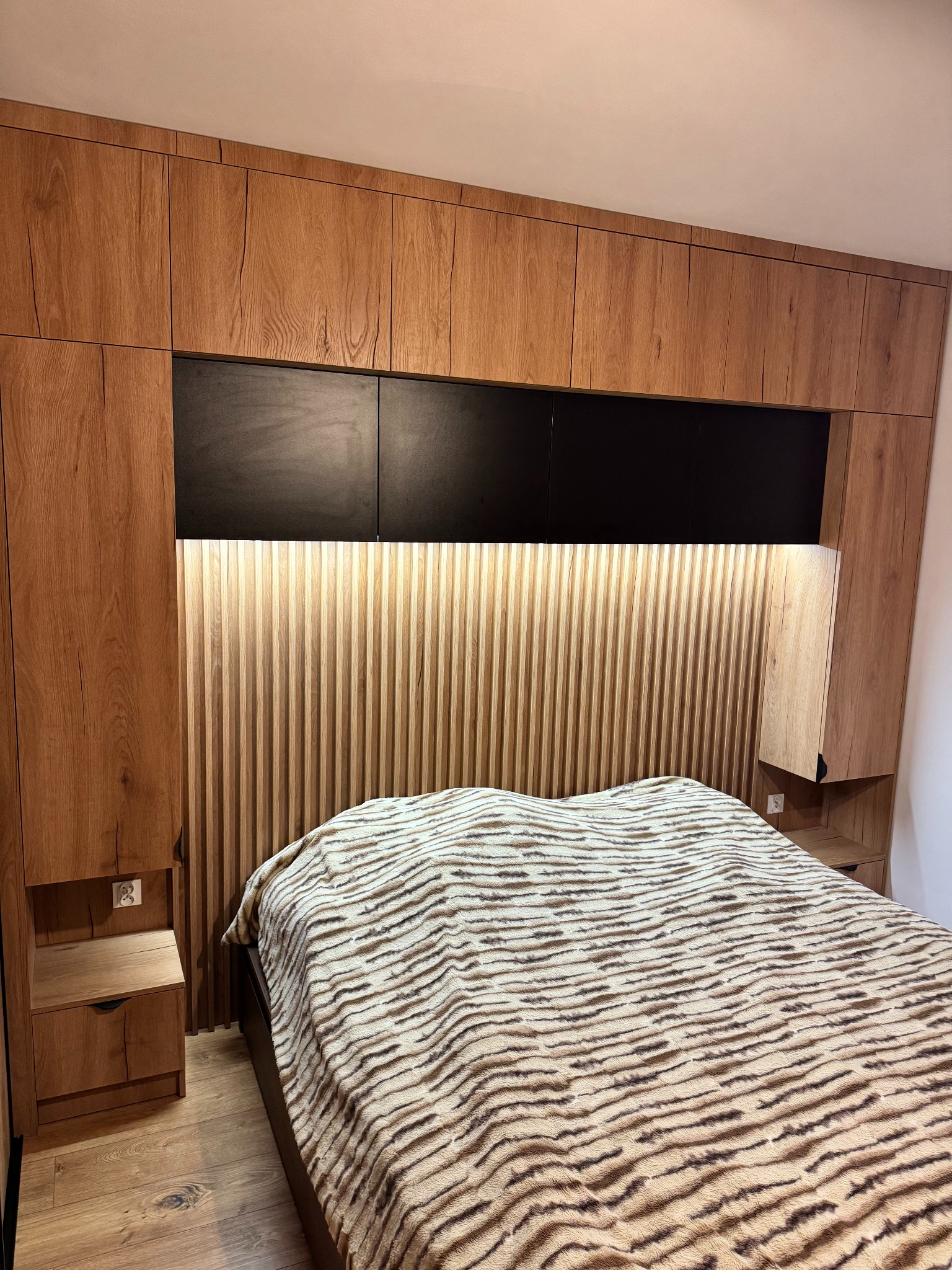 Drewniana zabudowa sypialni z szafkami nad łóżkiem i panelem dekoracyjnym z podświetleniem. Widoczne szafki nocne i wzorzysta narzuta na łóżku.