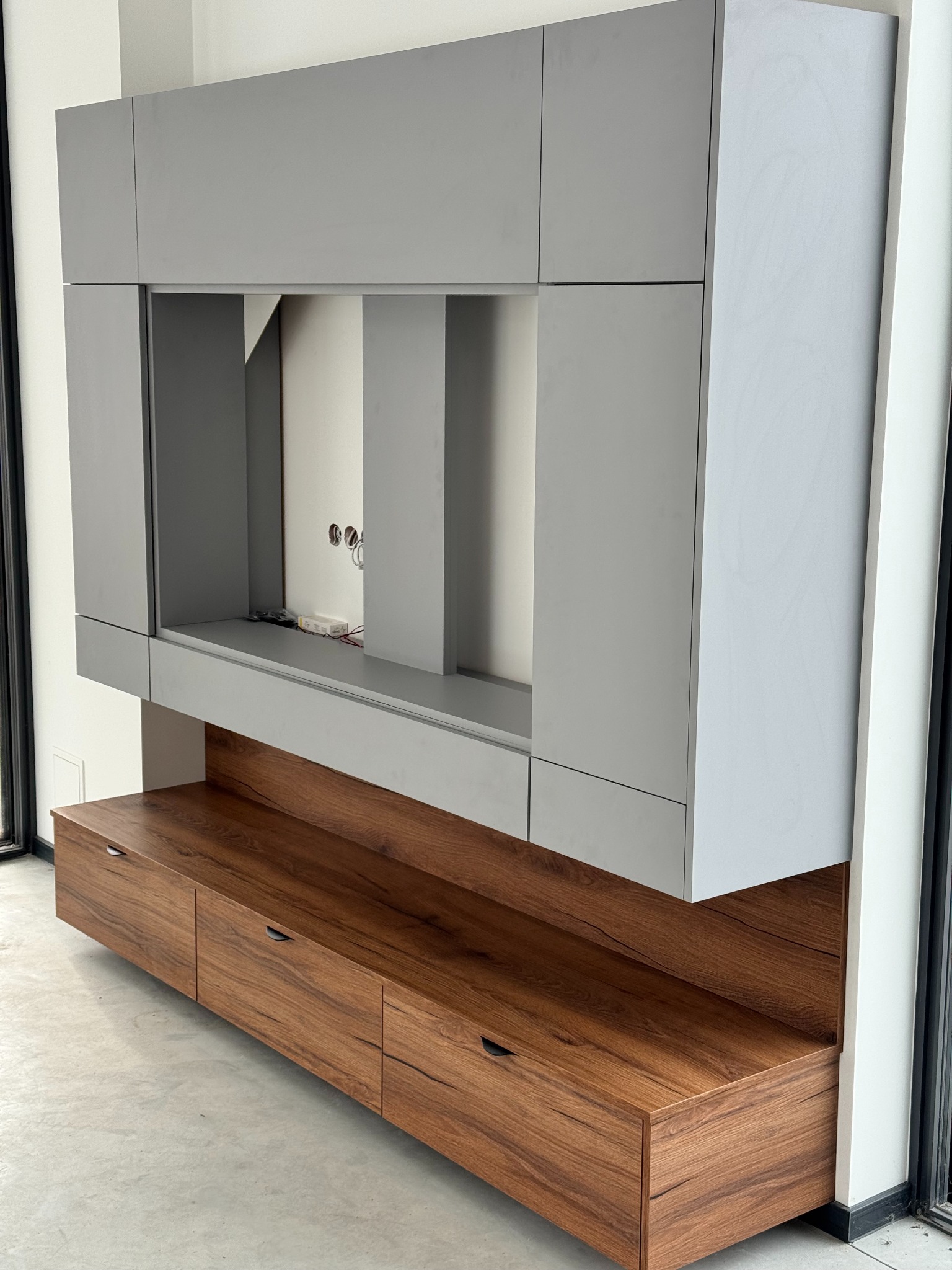 Szara zabudowa RTV z drewnianymi szufladami w nowoczesnym domu. Widoczne okno i surowy beton na podłodze. Minimalistyczny design.