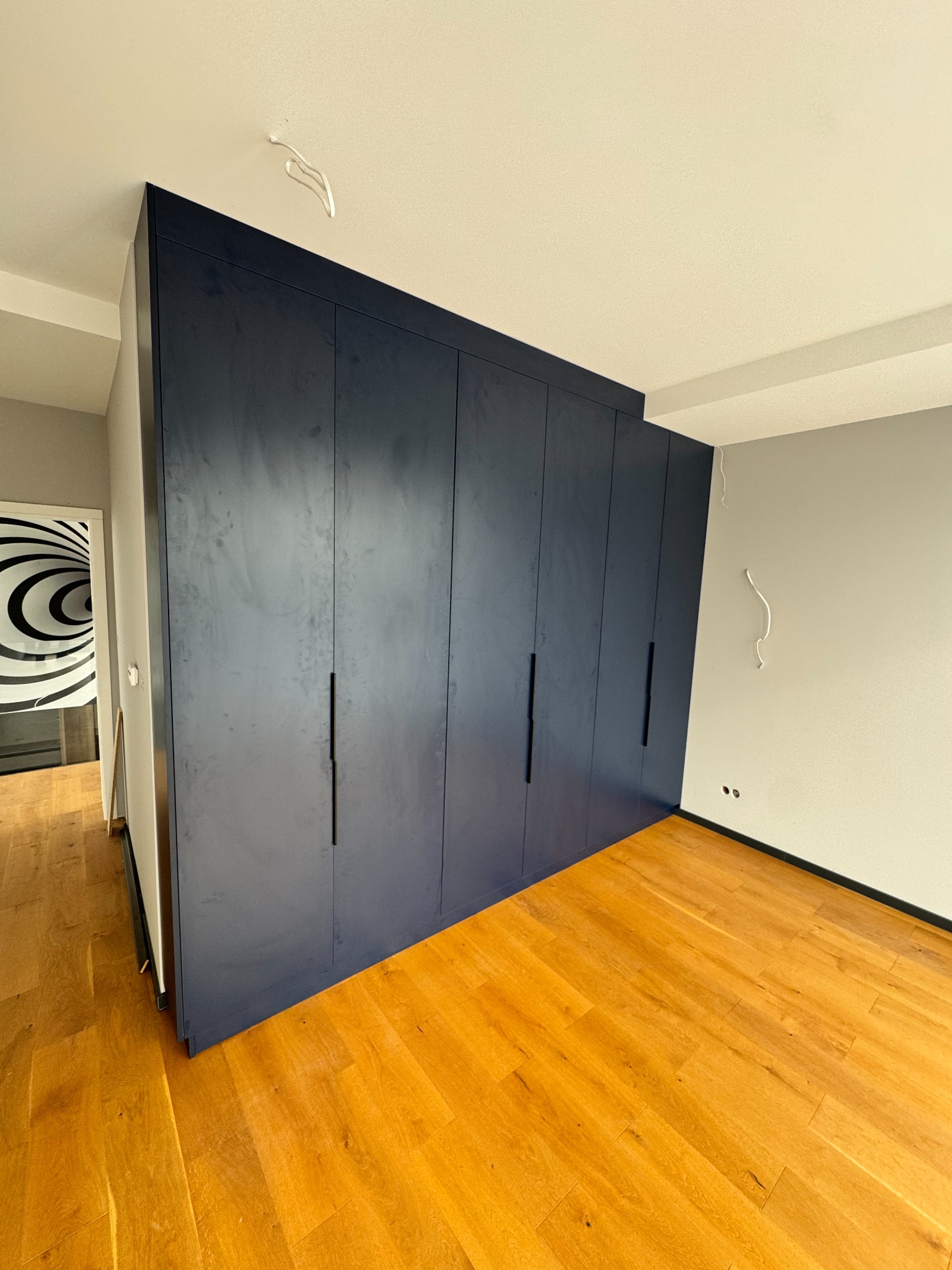 Granatowa szafa wnękowa z długimi, czarnymi uchwytami, na tle parkietu i szarej ściany. Widoczne wystające kable ze ściany i sufitu. W tle drzwi z abstrakcyjnym wzorem.