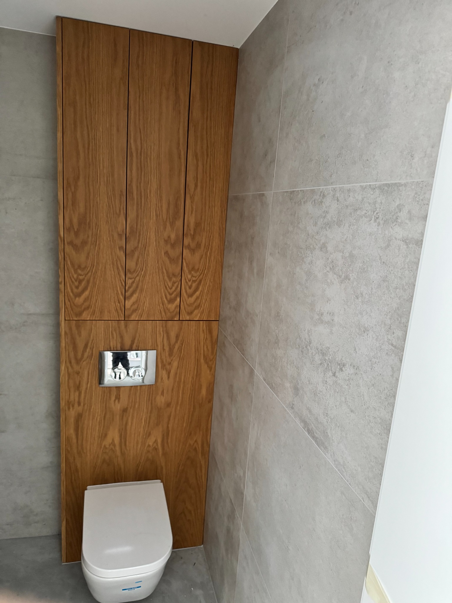 Drewniana zabudowa toalety z szafkami wiszącymi, szarymi płytkami i chromowanym przyciskiem spłukiwania. Minimalistyczny design, jasne wnętrze.