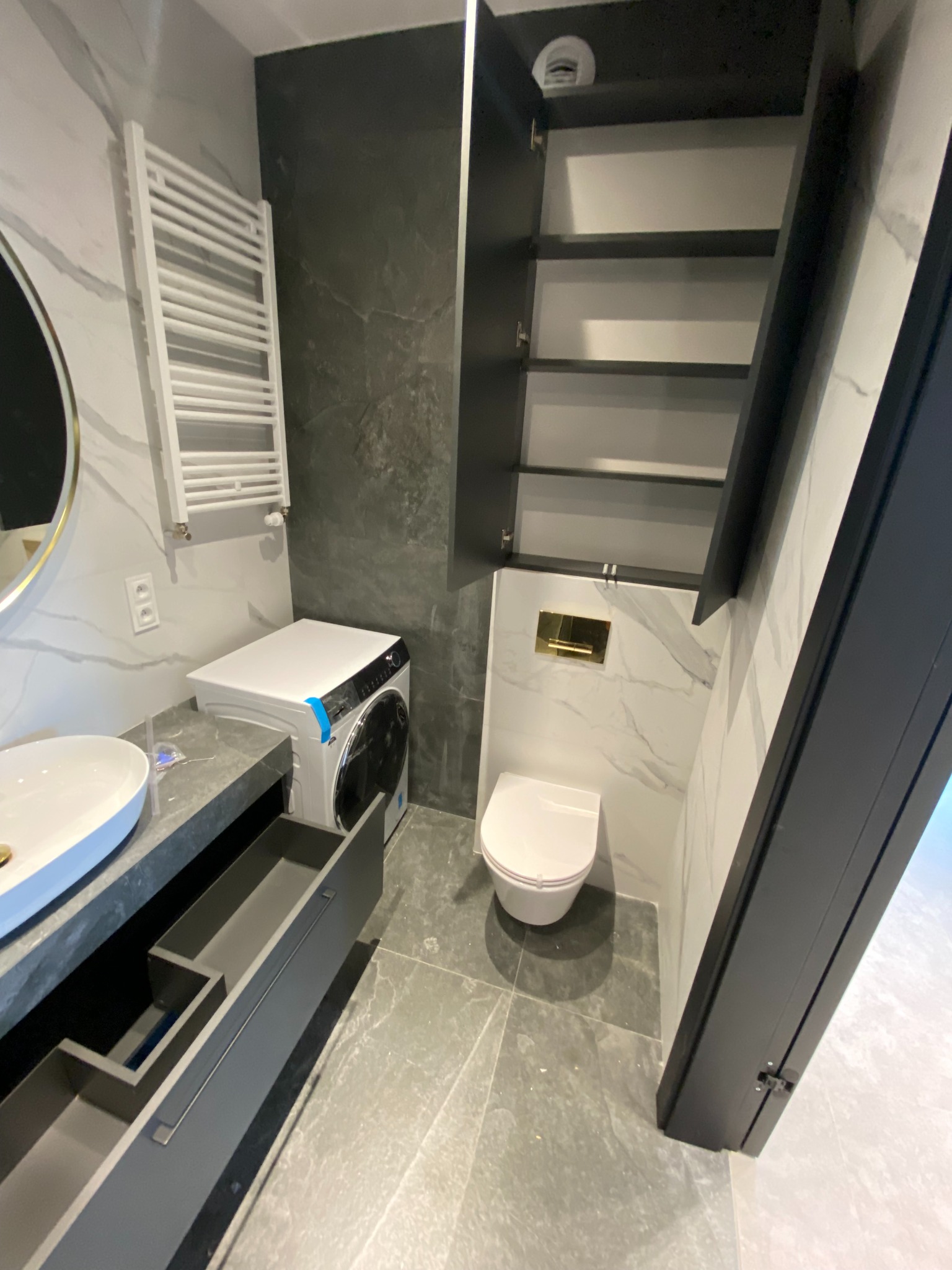 Nowoczesna łazienka z szarą szafką pod umywalką z otwartymi szufladami, pralką i podwieszaną szafką. Marmurowe płytki na ścianach i podłodze.