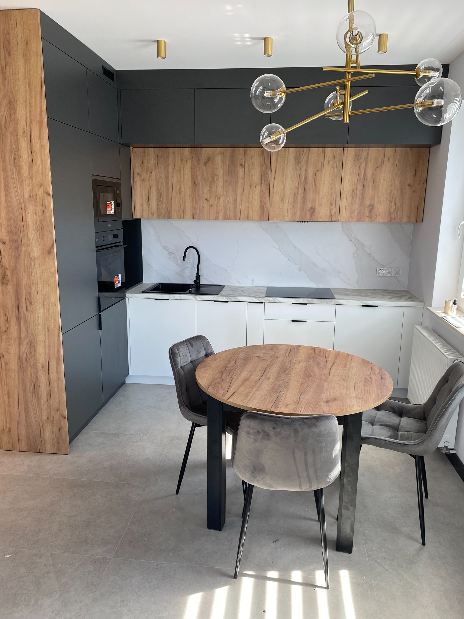 Nowoczesna kuchnia z drewnianymi i grafitowymi szafkami, okrągłym stołem i złotą lampą. Biały marmurkowy backsplash i szare krzesła dopełniają wystrój.