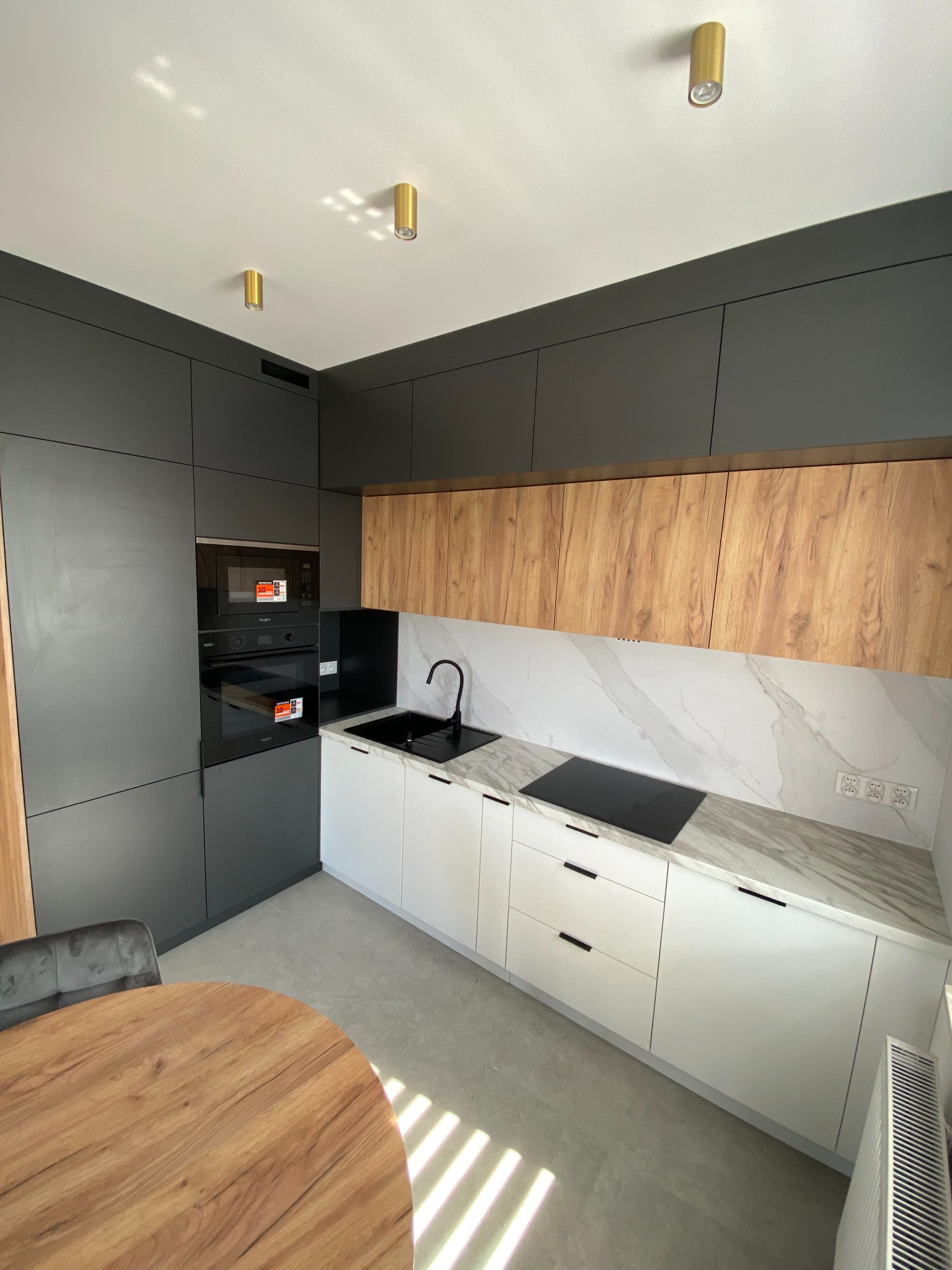 Nowoczesna kuchnia: grafitowe szafki, drewniane górne panele, białe dolne szafki z czarnymi uchwytami, marmurowy blat i okrągły drewniany stół.