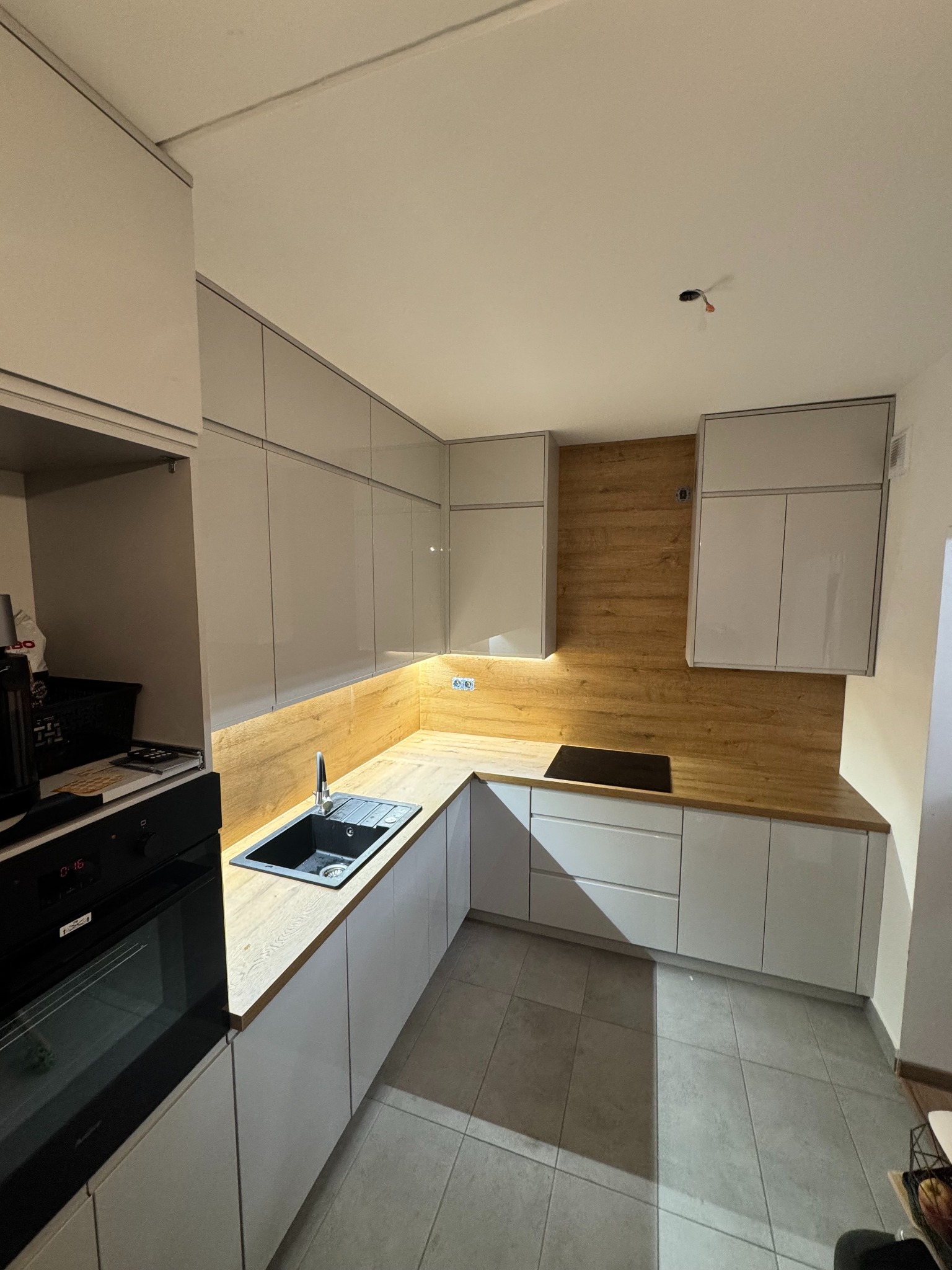 Jasna kuchnia z drewnianym blatem i czarnym zlewem. Górne szafki lakierowane na wysoki połysk. Widoczny fragment piekarnika i płyty grzewczej. Oświetlenie LED pod szafkami.