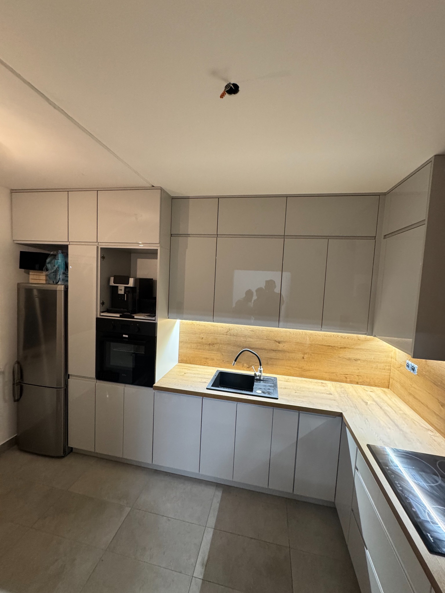 Nowoczesna kuchnia na wymiar z białymi, lakierowanymi frontami, drewnianym blatem i oświetleniem LED pod szafkami wiszącymi. Widoczny zlew, płyta indukcyjna i zabudowany sprzęt.