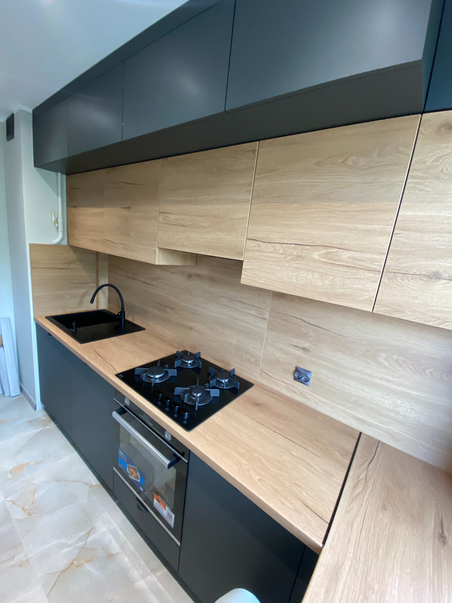Nowoczesna kuchnia z drewnianymi frontami szafek i czarnymi elementami: zlew, bateria, płyta grzewcza i szafki. Jasna podłoga z marmurowym wzorem.