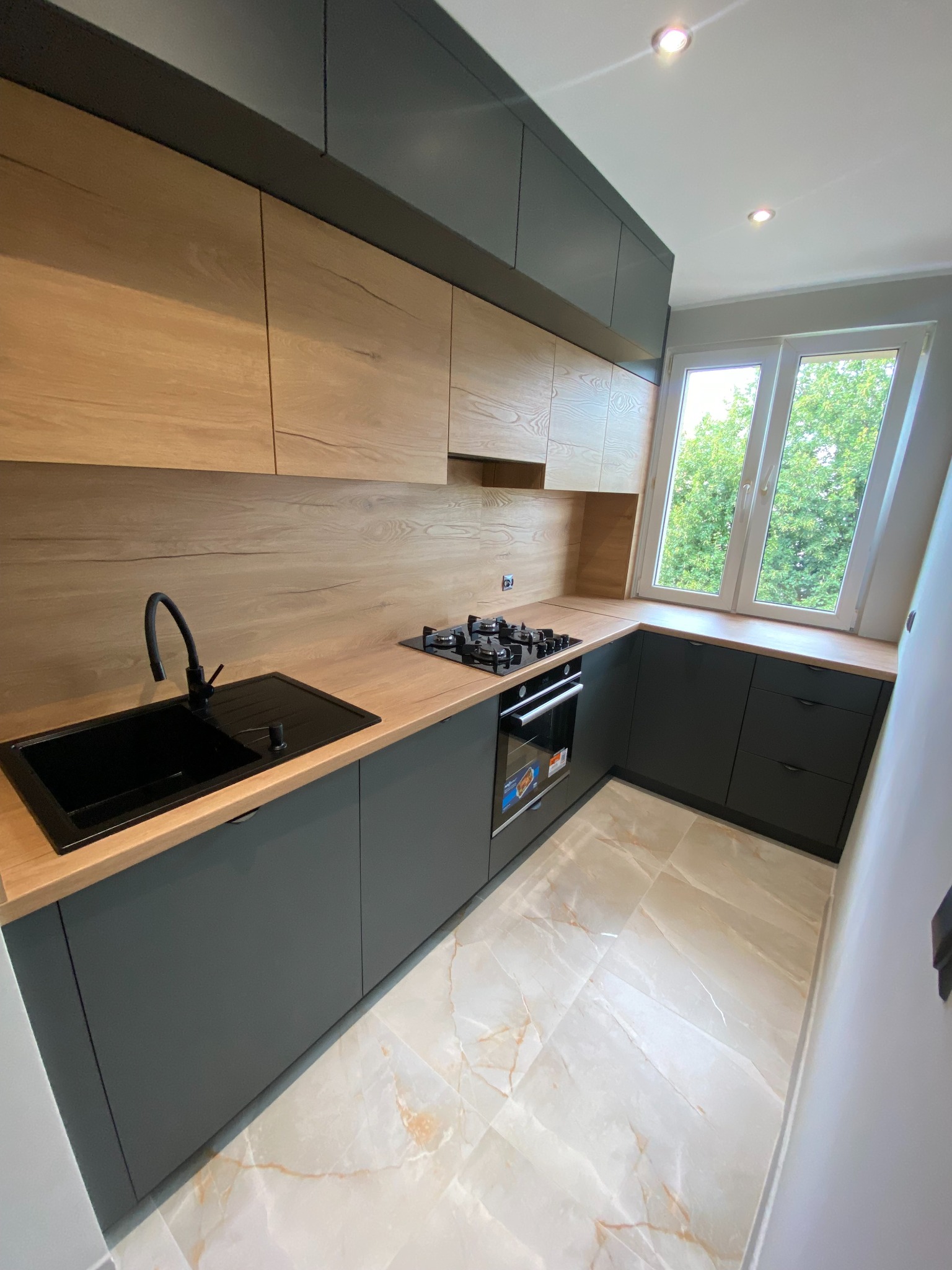 Nowoczesna kuchnia na wymiar z drewnianymi frontami szafek górnych i grafitowymi dolnymi. Czarny zlew i bateria, płyta gazowa przy oknie. Marmurowe płytki podłogowe.