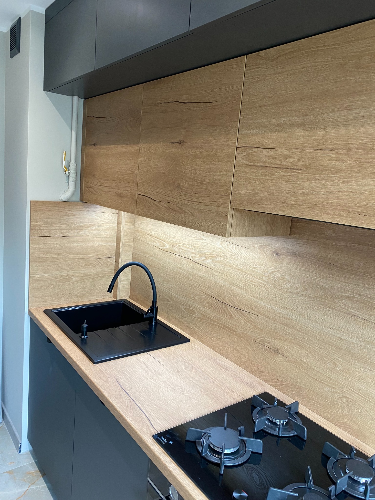 Nowoczesna kuchnia z drewnianymi frontami szafek i czarnym blatem, z widocznym zlewem i płytą grzewczą. Oświetlenie LED pod szafkami górnymi.
