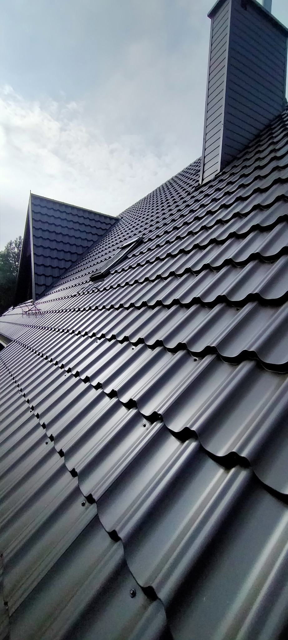 Nowe, grafitowe pokrycie dachowe z blachy trapezowej, widoczne okno dachowe i komin na tle pochmurnego nieba.