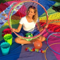 Uśmiechnięta kobieta w okularach przeciwsłonecznych, siedząca na kolorowej macie, trzymająca dwa koła hula-hop, z piłką i kolorowymi wiaderkami wokół.
