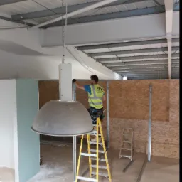 Pracownik w kamizelce odblaskowej na żółtej drabinie montuje instalację wewnątrz budynku z widoczną konstrukcją sufitu i ścianami z płyty OSB oraz częściowo zamontowaną płytą kartonowo-gipsową.
