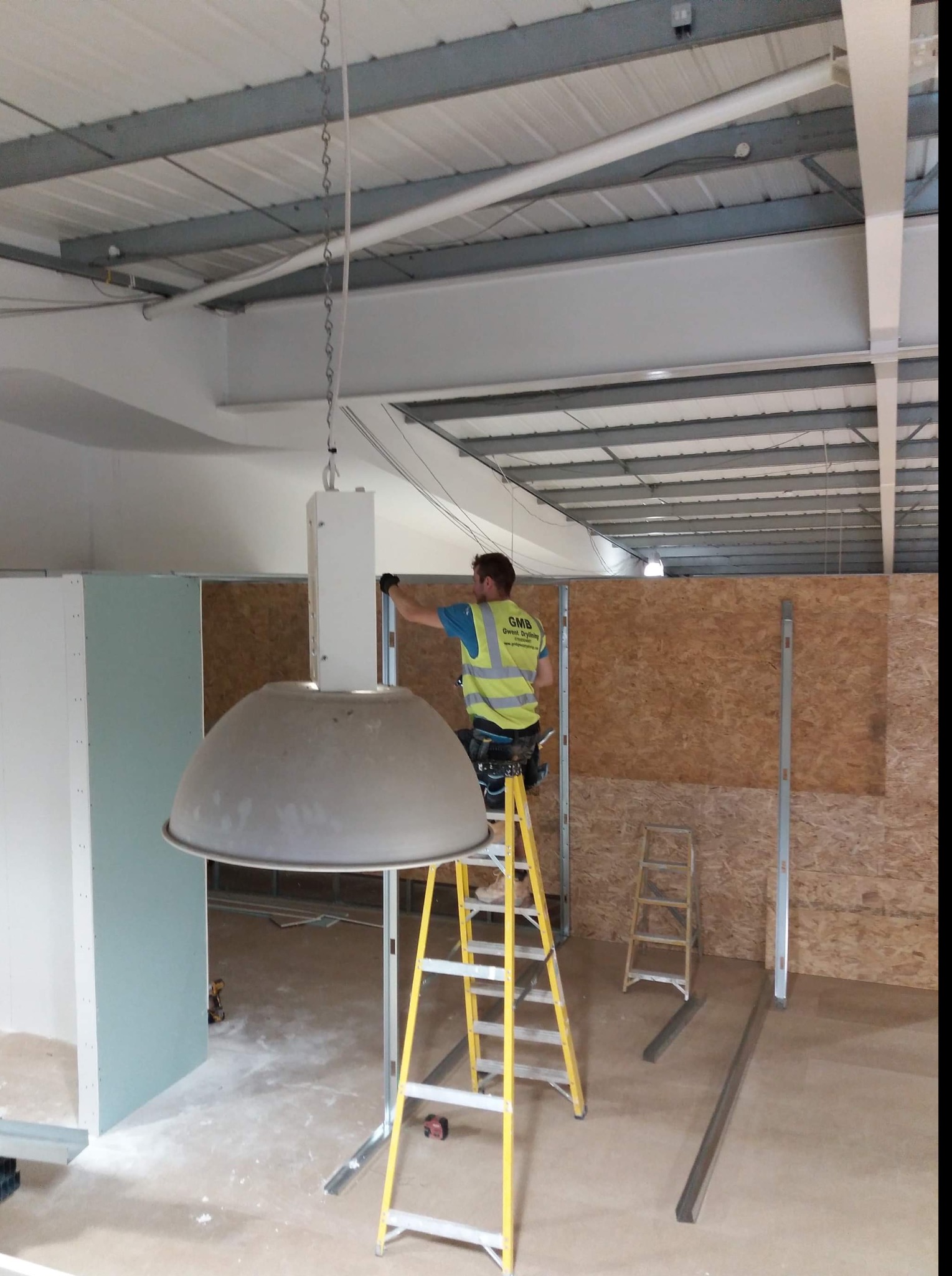 Pracownik w kamizelce odblaskowej na żółtej drabinie montuje instalację wewnątrz budynku z widoczną konstrukcją sufitu i ścianami z płyty OSB oraz częściowo zamontowaną płytą kartonowo-gipsową.