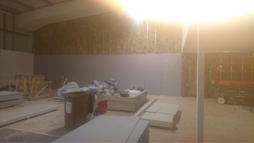 Wnętrze w trakcie remontu: widoczne płyty gipsowo-kartonowe przygotowane do montażu, metalowe profile na ścianie z izolacją, podnośnik nożycowy w tle, materiały budowlane i śmietnik na podłodze.