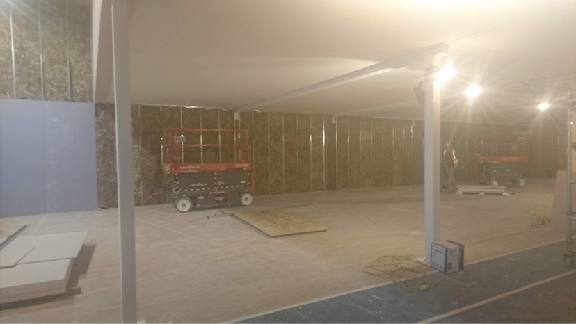 Wnętrze w trakcie prac wykończeniowych: metalowe profile na ścianach z izolacją, płyta kartonowo-gipsowa oparta o ścianę, podnośnik nożycowy Skyjack i spawarka na podłodze.