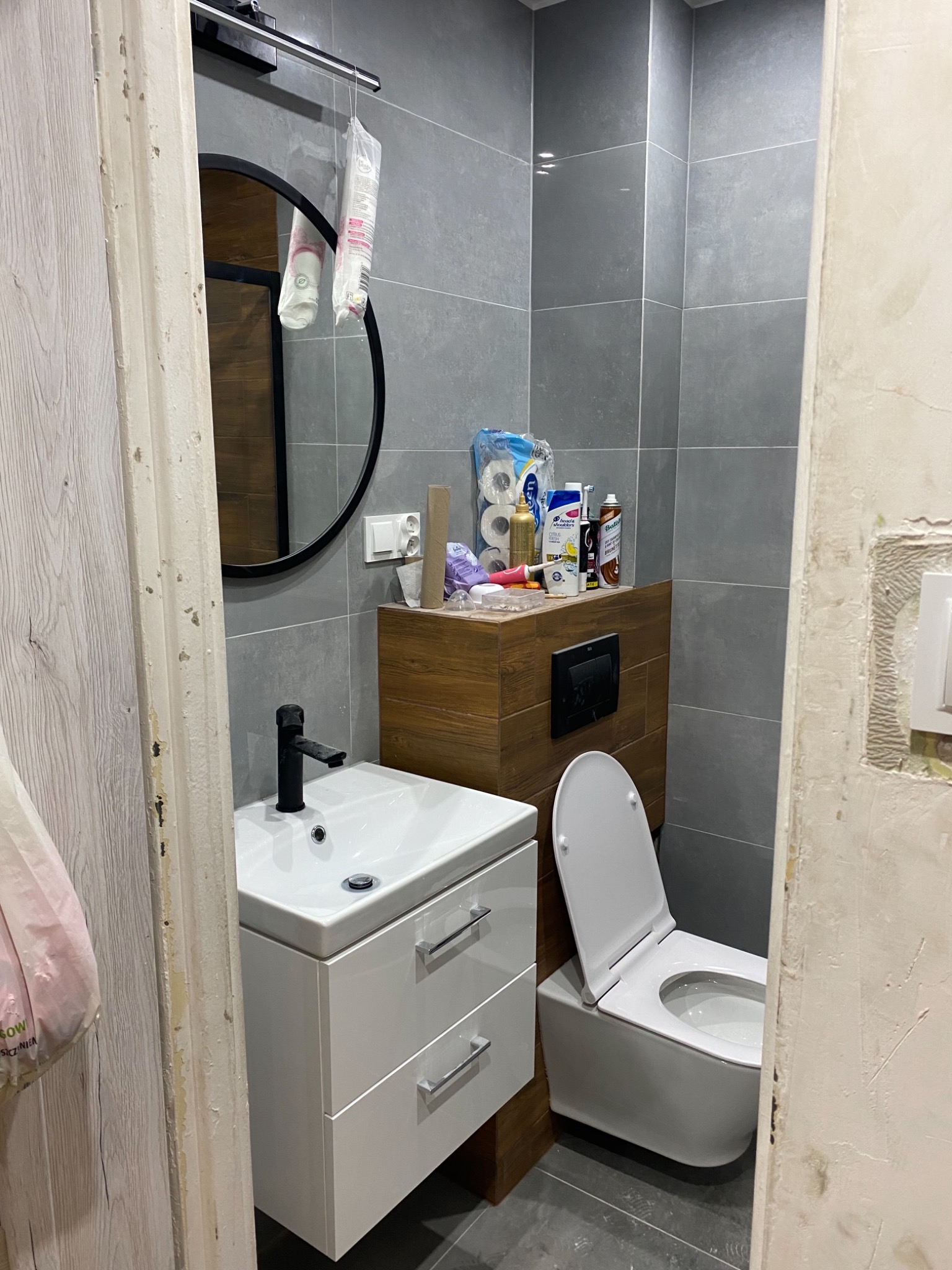 Wnętrze łazienki w trakcie remontu, widoczna umywalka z szafką, toaleta oraz fragment ściany z szarymi płytkami i fragment ściany przygotowanej do malowania. Widoczne niedoskonałości na framudze...