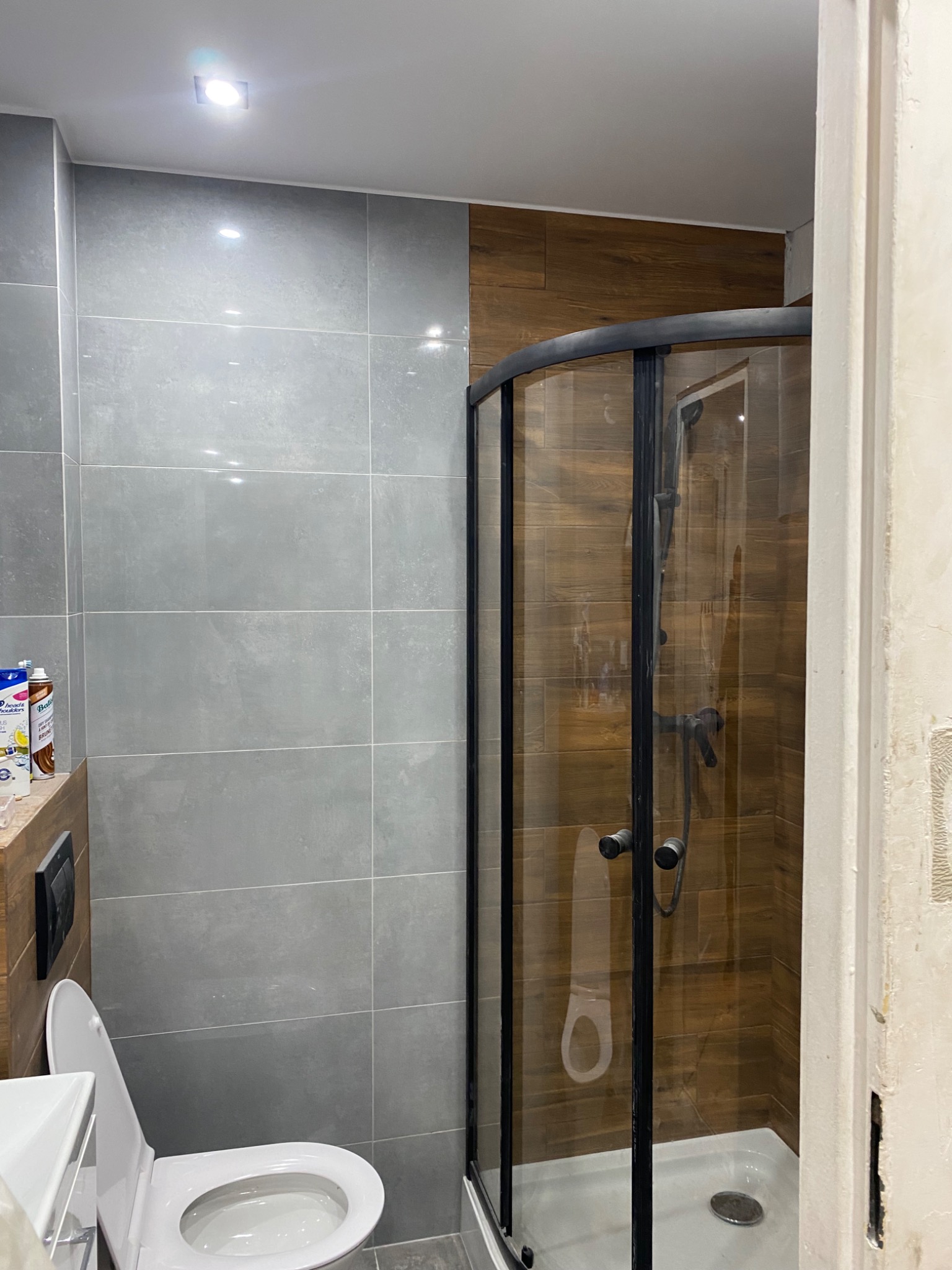 Nowoczesna łazienka po remoncie: szare płytki ścienne, kabina prysznicowa z czarnymi profilami i drewnopodobne płytki wewnątrz kabiny, toaleta z otwartą klapą.
