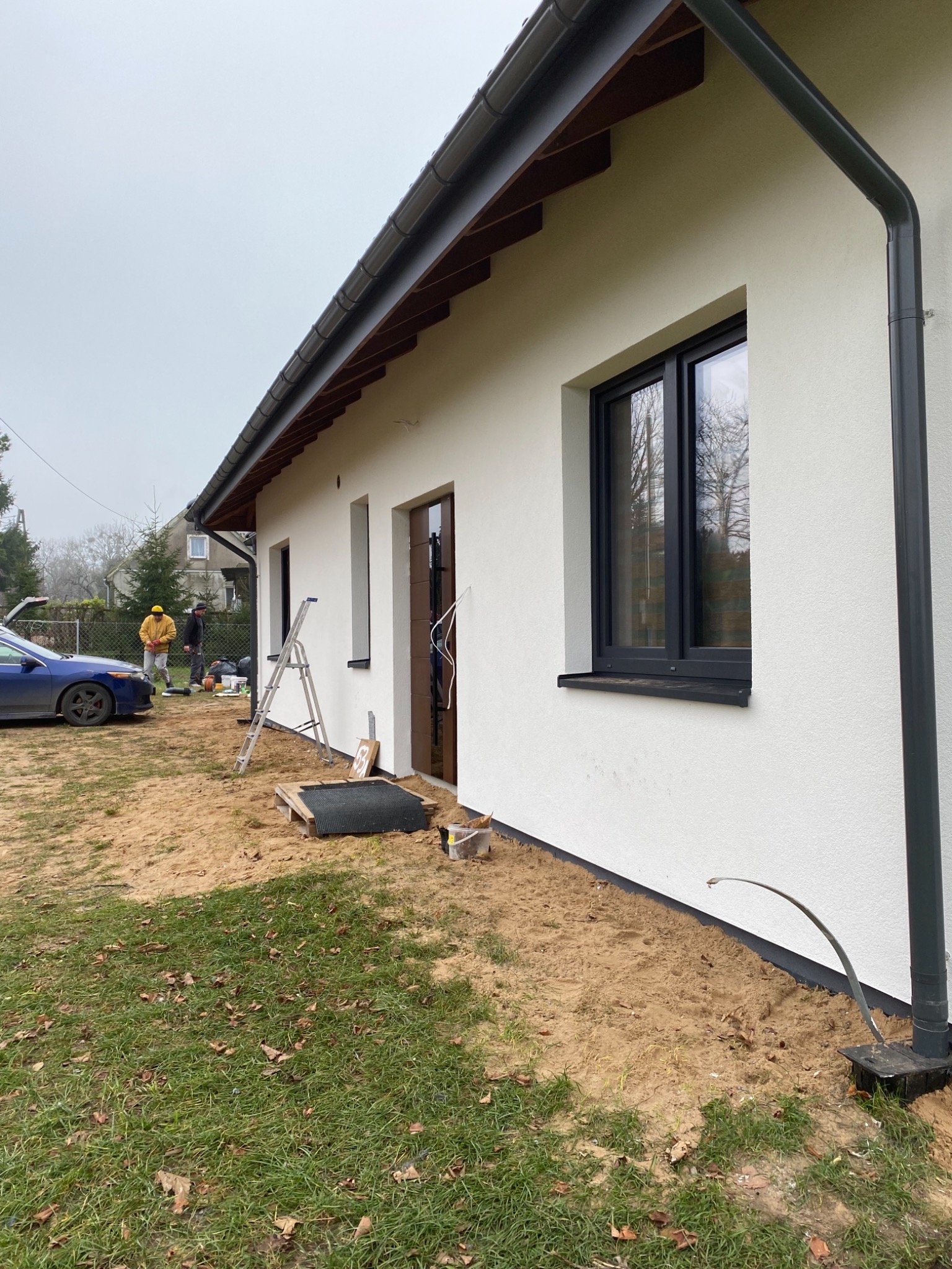 Boczny widok domu z nową białą elewacją, ciemnymi oknami i orynnowaniem, z pracownikami w tle i samochodem zaparkowanym obok, na trawniku z piaskiem i liśćmi.