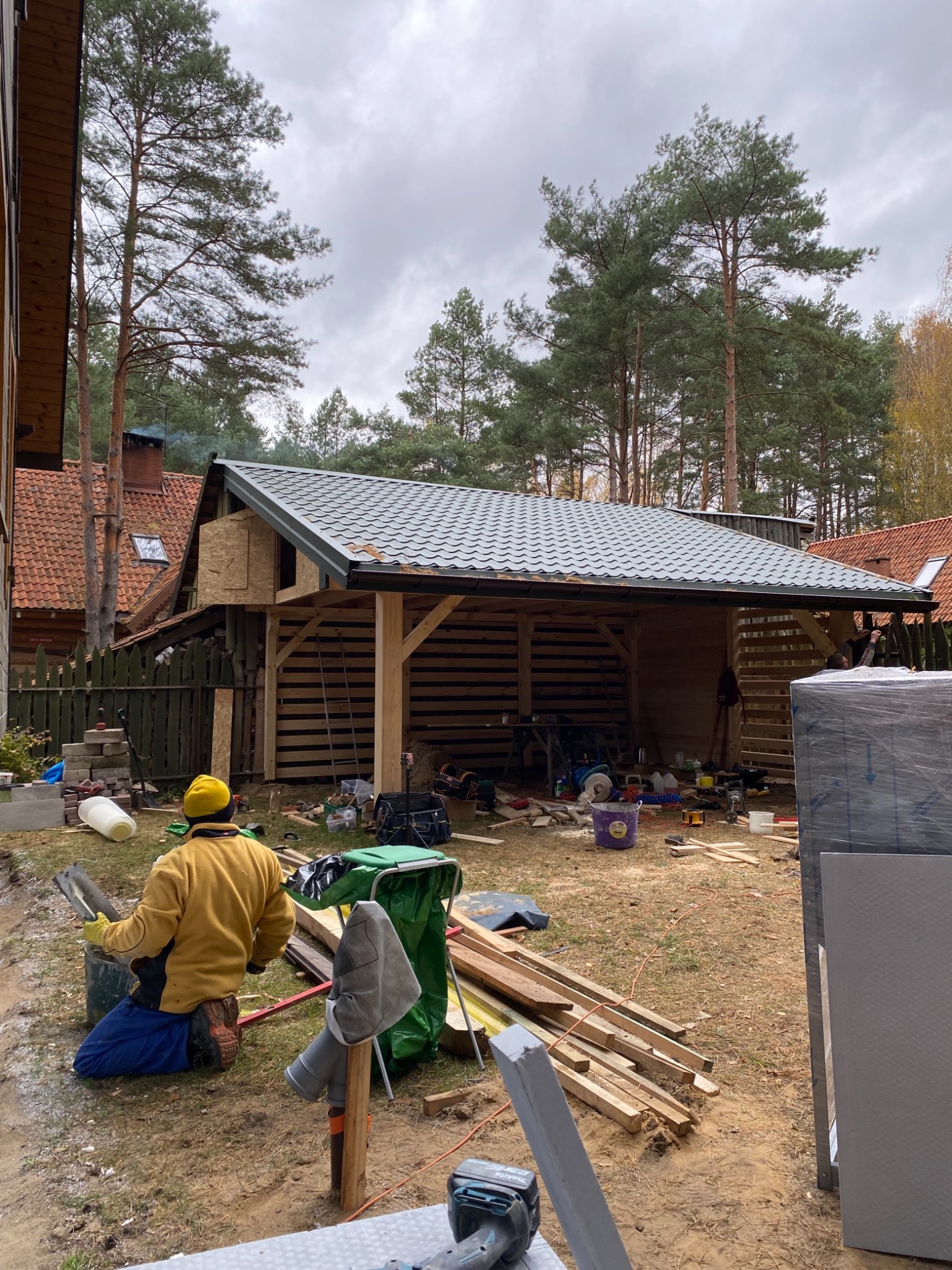 Budowa drewnianej wiaty z szarą blachodachówką w ogrodzie, widok zza pracownika w żółtej kurtce, narzędzia i materiały budowlane porozrzucane wokół.