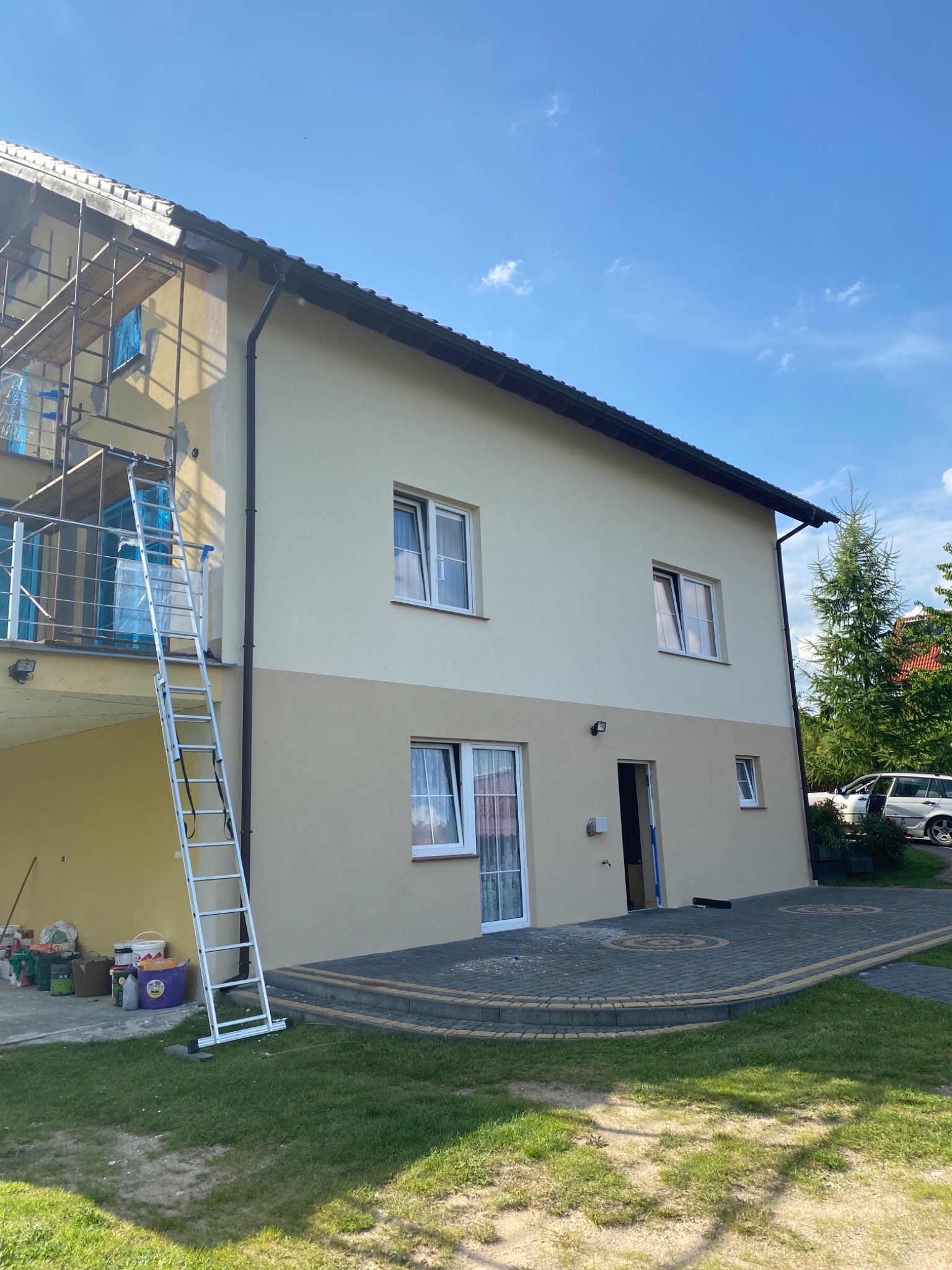Dwukondygnacyjny dom po częściowym odnowieniu elewacji, z widocznymi rusztowaniami i drabiną, świeżo pomalowany górny fragment w odcieniach beżu, dolny w kolorze piaskowym, na trawniku widoczne...
