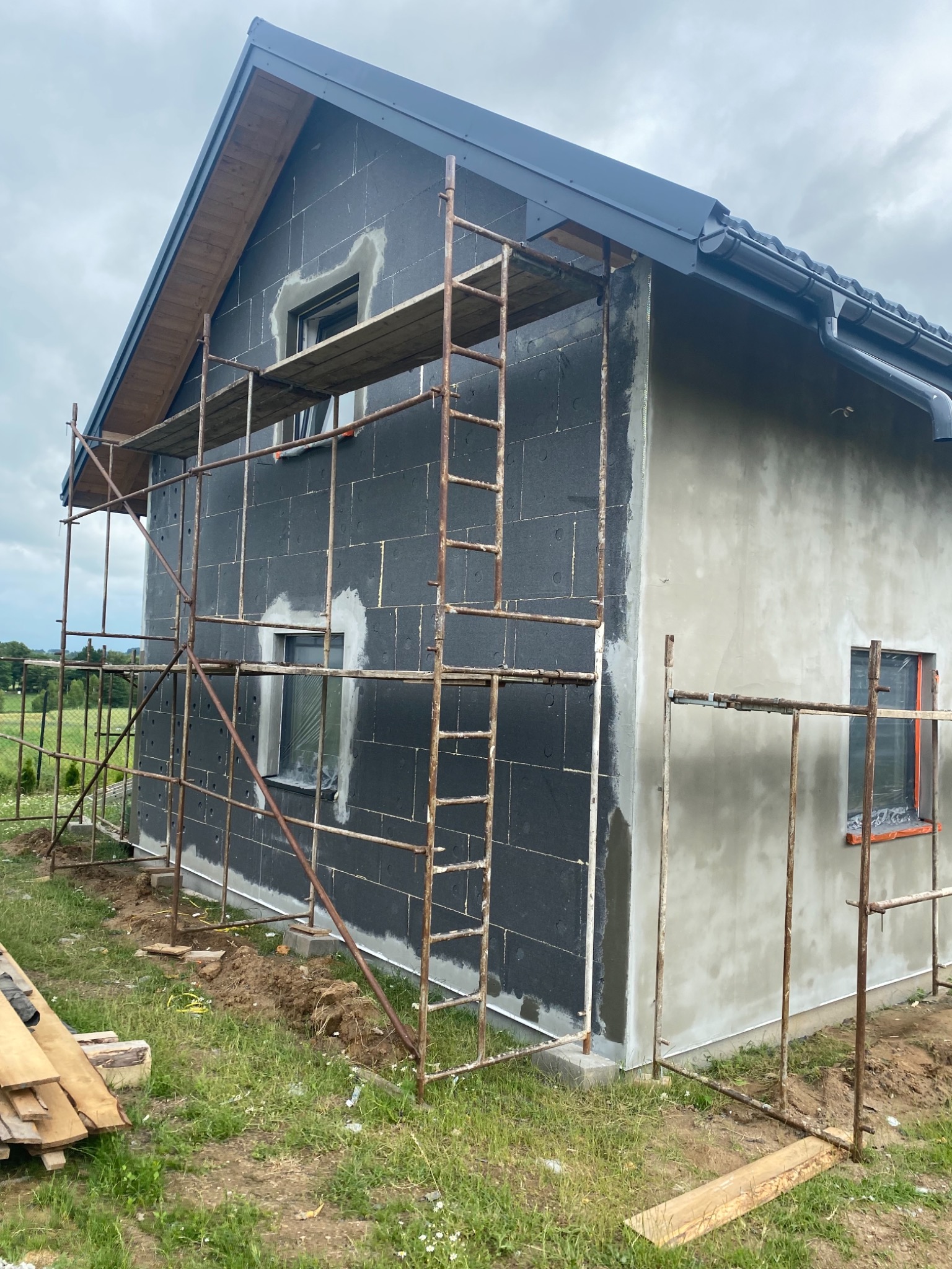 Dom w trakcie ocieplania: czarny styropian na elewacji, fragment otynkowanej ściany, rusztowanie metalowe, drewniane deski obok.