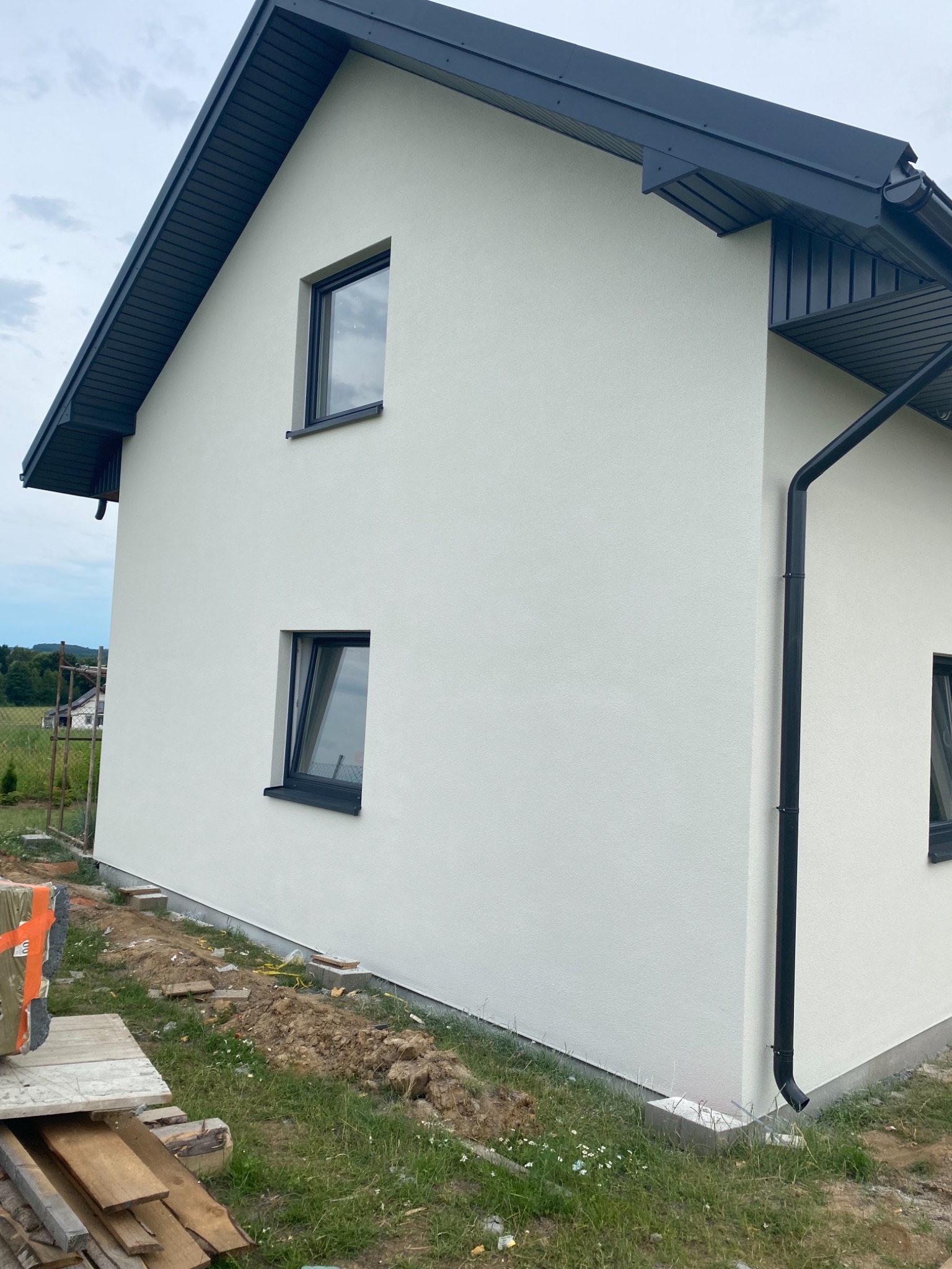 Świeżo otynkowana elewacja domu jednorodzinnego w kolorze jasnej bieli, z czarnymi oknami i rynnami, widok z zewnątrz na narożnik budynku w trakcie prac wykończeniowych.