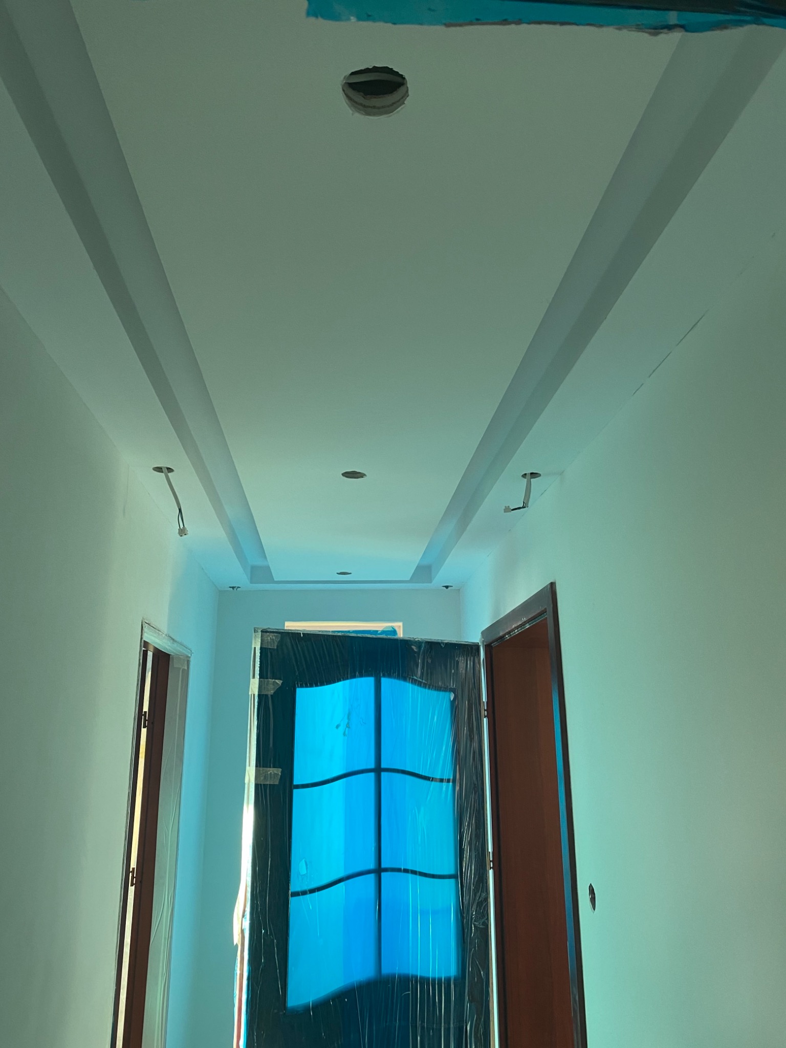 Wnętrze w trakcie prac wykończeniowych: widok na sufit z listwami dekoracyjnymi, otwory na oświetlenie oraz drzwi zabezpieczone folią ochronną.