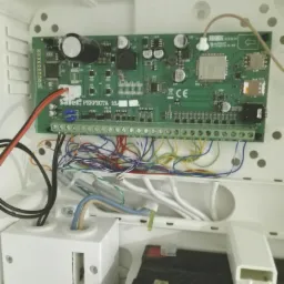 Wnętrze obudowy centrali alarmowej Satel Perfecta 16 z widoczną płytą główną, licznymi przewodami podłączonymi do listwy zaciskowej, akumulatorem i elementami montażowymi.