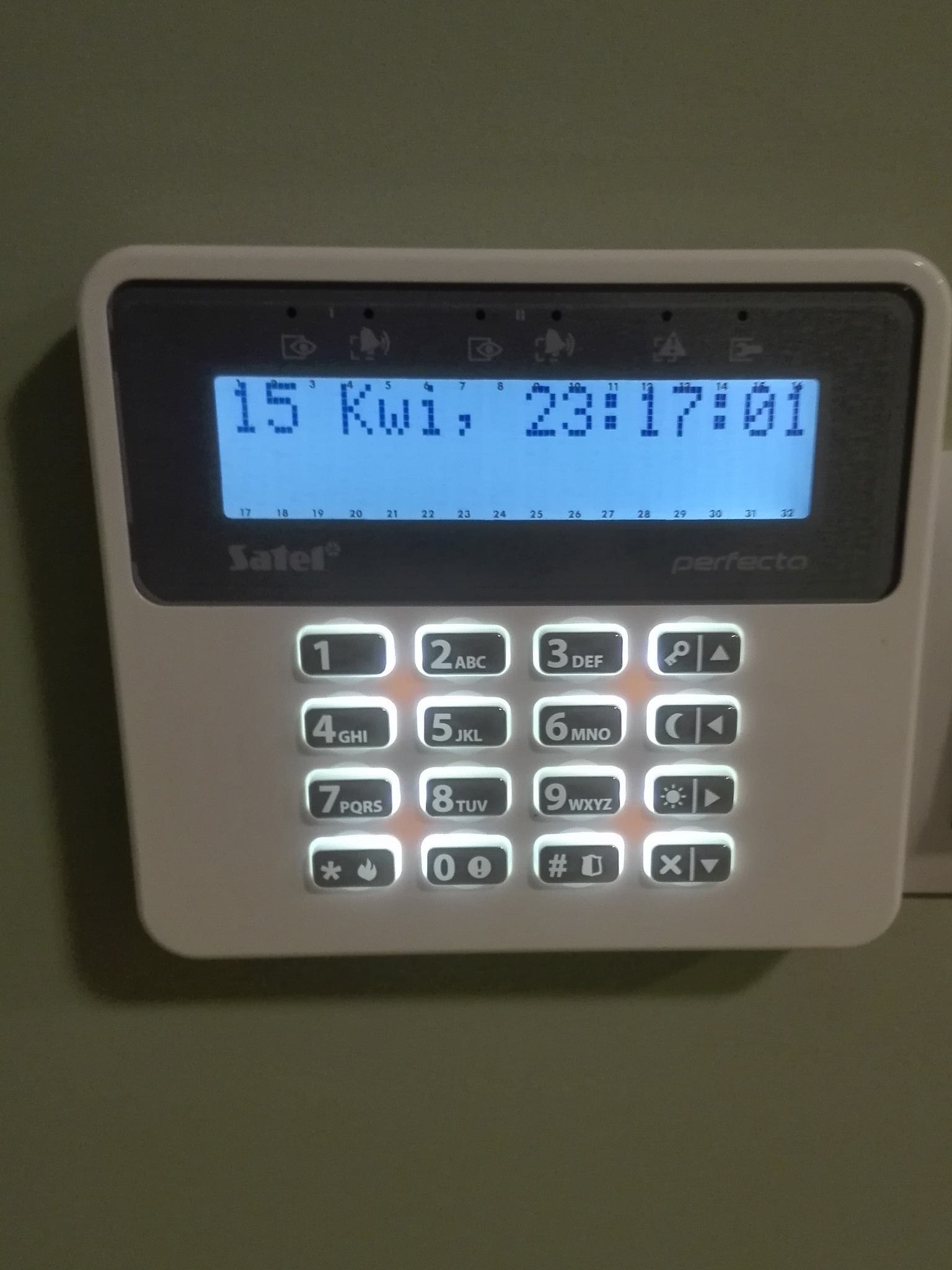 Panel sterowania alarmu domowego Satel Perfecta z podświetlanymi przyciskami, wyświetlający datę 15 kwietnia i godzinę 23:17:01, zamontowany na zielonej ścianie.