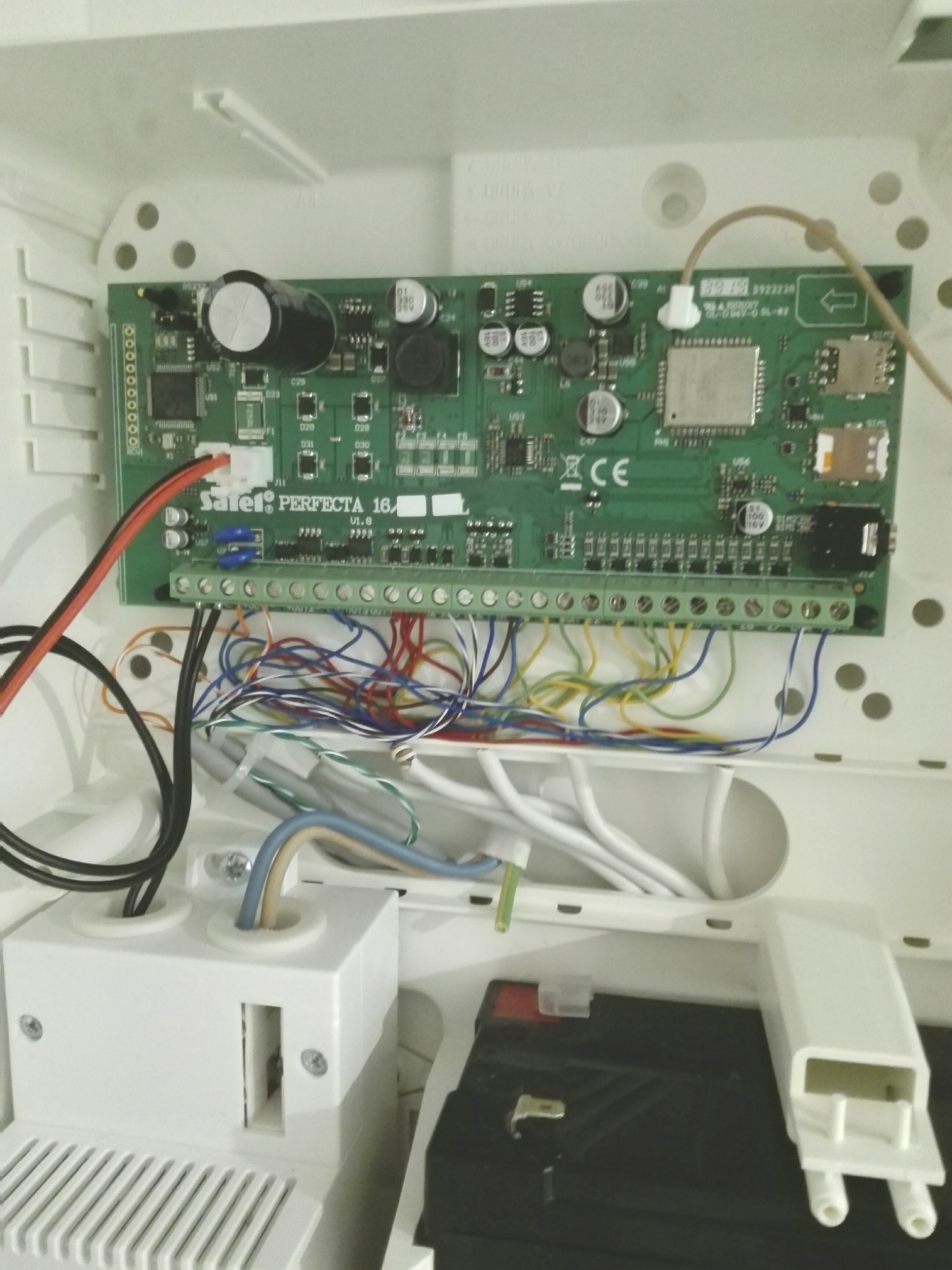 Wnętrze obudowy centrali alarmowej Satel Perfecta 16 z widoczną płytą główną, licznymi przewodami podłączonymi do listwy zaciskowej, akumulatorem i elementami montażowymi.
