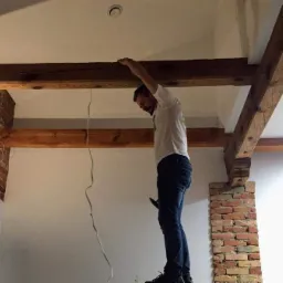 Elektryk na drabinie, sprawdza instalację elektryczną pod sufitem z drewnianymi belkami w pomieszczeniu z ceglanymi elementami dekoracyjnymi.