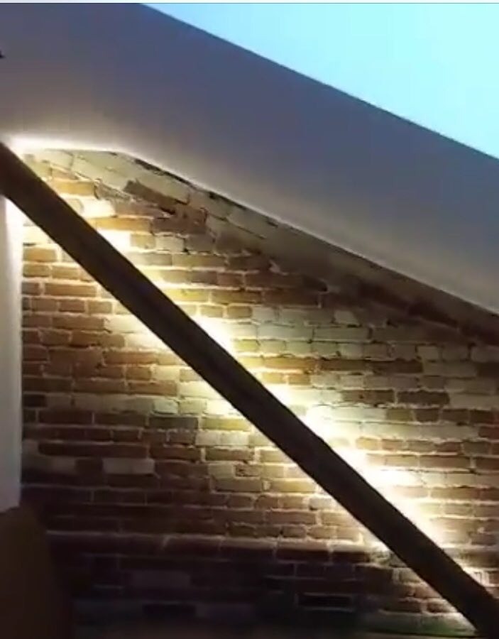 Podświetlona ceglana ściana w poddaszu z drewnianą belką, oświetlenie punktowe skierowane ku górze.