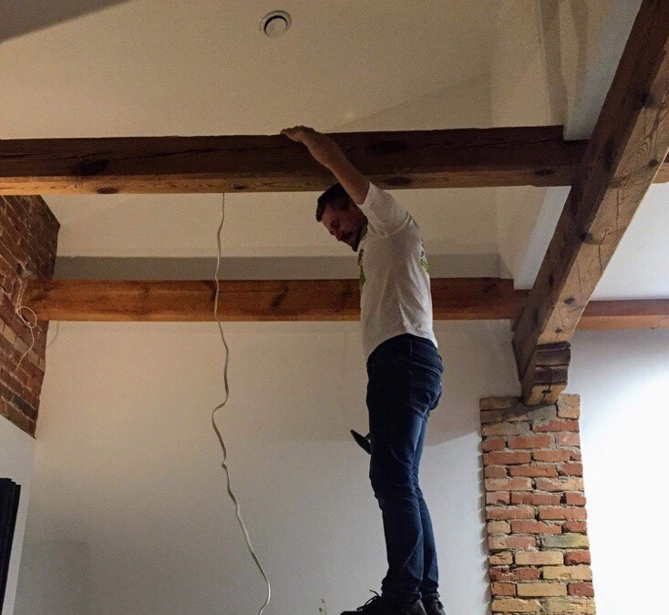 Elektryk na drabinie, sprawdza instalację elektryczną pod sufitem z drewnianymi belkami w pomieszczeniu z ceglanymi elementami dekoracyjnymi.