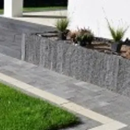 Nowoczesne schody kamienne prowadzące do domu w Obornikach, z szarej kostki, otoczone zielonym trawnikiem i rabatą z roślinami ozdobnymi. Widoczny fragment elewacji budynku.