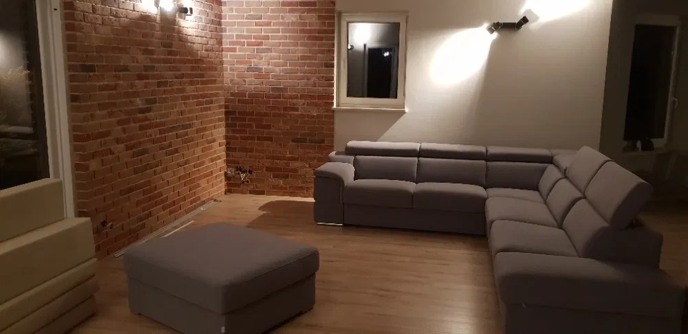 Ściana z cegły klinkierowej w salonie, szara narożna sofa z regulowanymi zagłówkami, oświetlenie punktowe na ścianach, przygotowana do wykończenia.