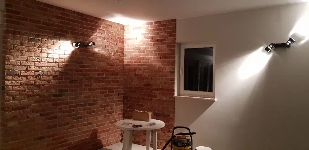 Wykończone wnętrze z ceglaną ścianą, oświetleniem punktowym i widocznym stolikiem z akcesoriami oraz odkurzaczem, sugerujące prace wykończeniowe.