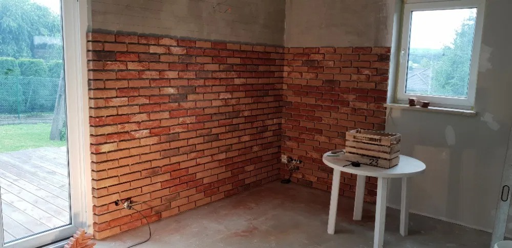Ściana wyłożona cegłą klinkierową w trakcie prac wykończeniowych, widoczne niedomalowane fragmenty ściany, mały biały stolik z drewnianą skrzynką i okno z widokiem na ogród.